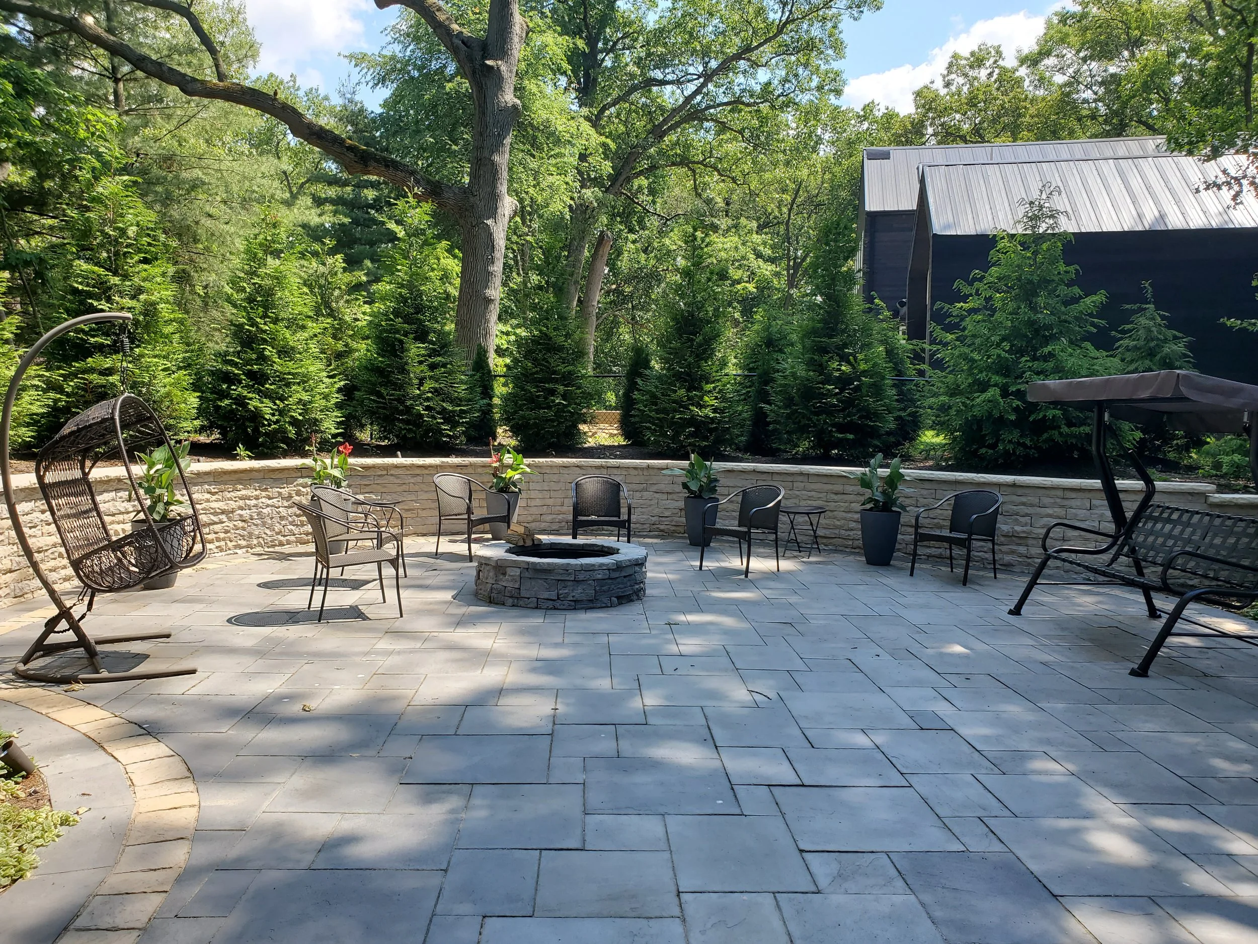An outdoor patio area with various chairs, a stone fire pit in the center, and a hanging swing chair on the left. The patio is surrounded by lush green trees and shrubs, with a stone wall in the background. There is a black structure, possibly a barn or shed, on the right side.