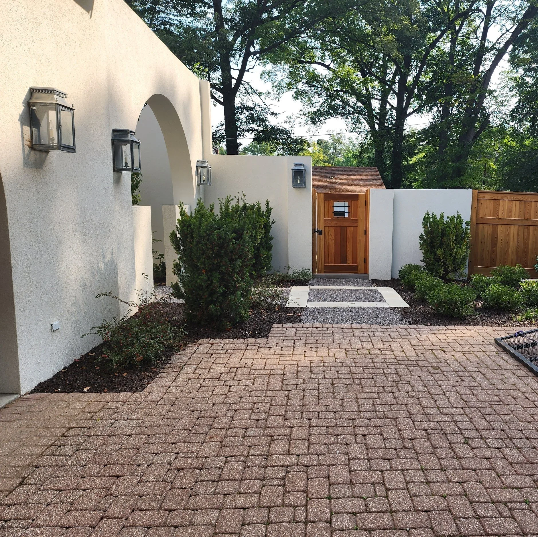 A courtyard with a paved brick surface, a white stucco wall with outdoor lights, a wooden gate, and green shrubs, with trees in the background.