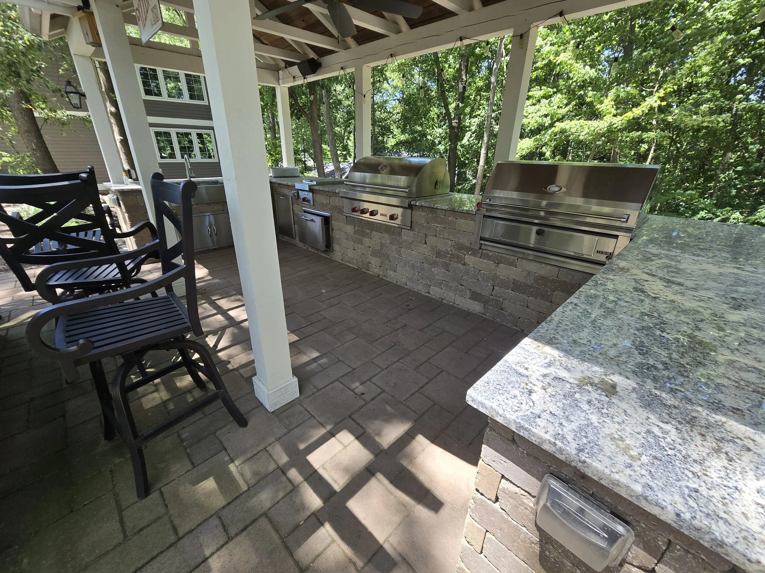 Outdoor kitchen with stainless steel grills, brick counter, granite prep station, and black chairs under a wooden roof, surrounded by trees.