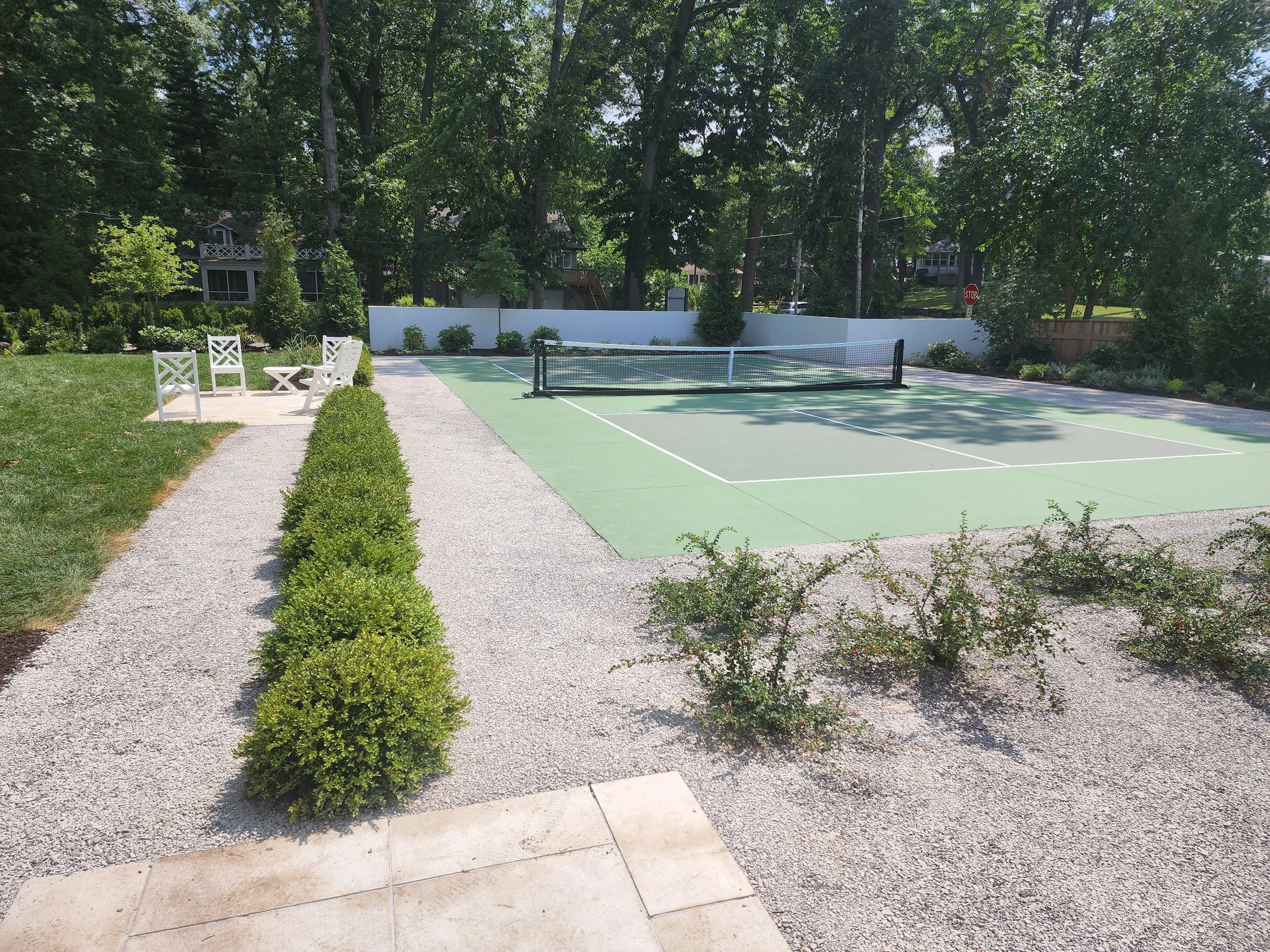 A fenced tennis court with a net, surrounded by trees and plants, with a white wall and some seating area with three white chairs and a small table on a gravel path.