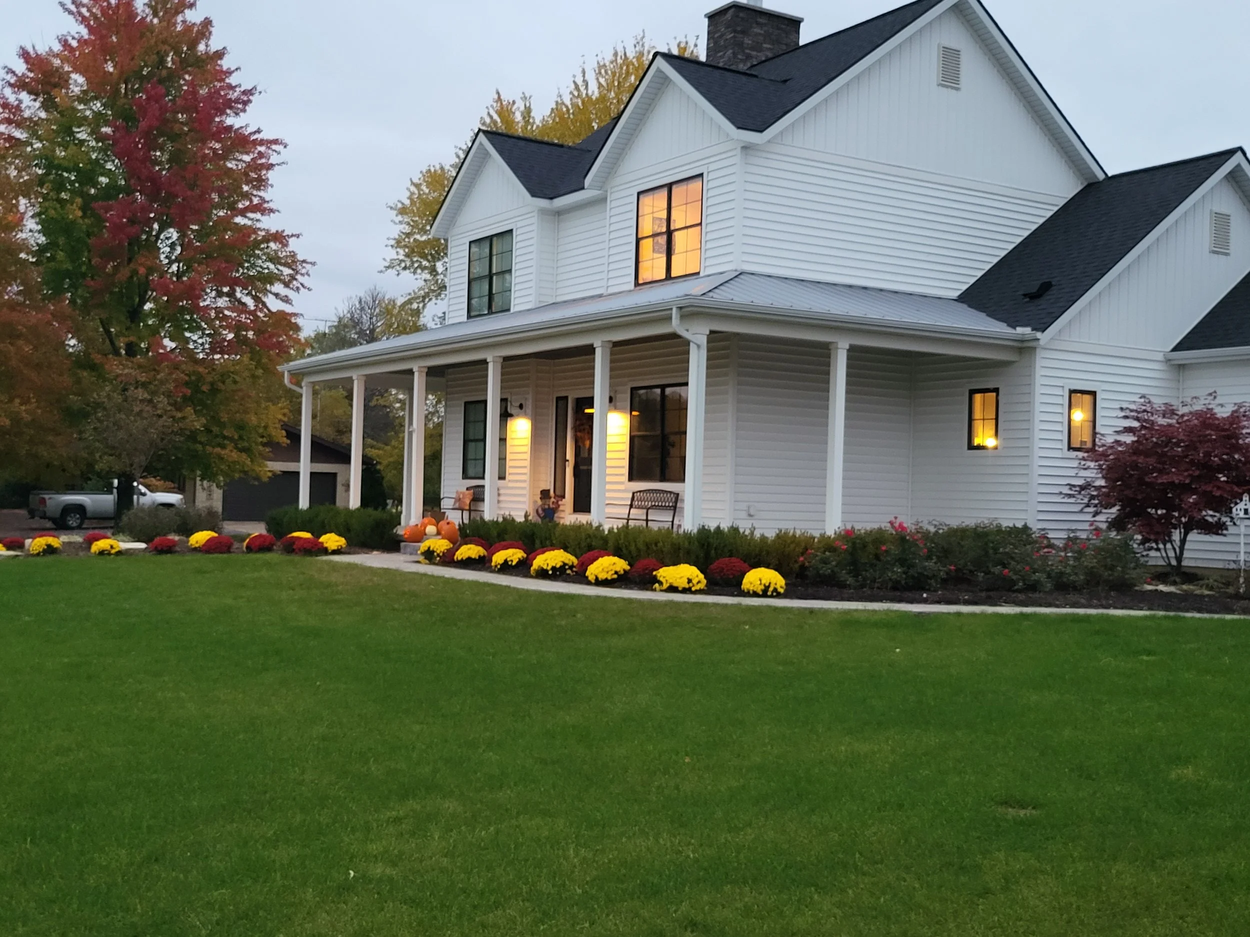 A white two-story house with a porch, surrounded by well-maintained lawn and fall flowers, with trees showing autumn colors in the background, and lights turned on inside as the evening approaches.