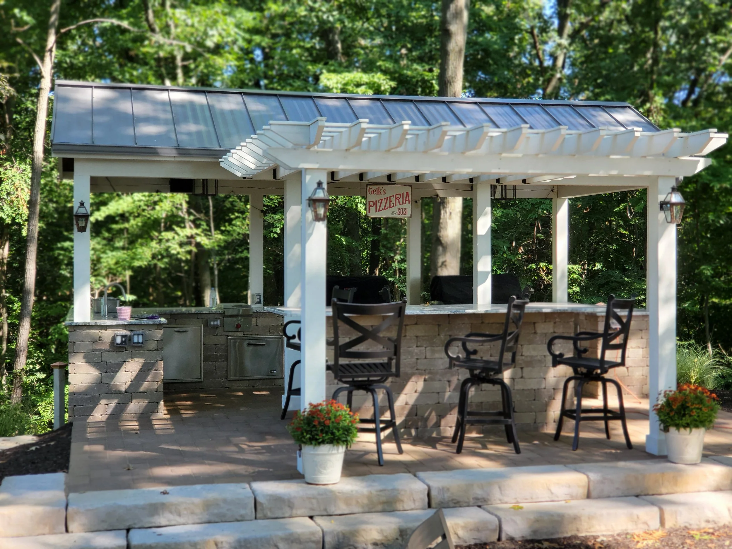 An outdoor bar with a brick counter, four black bar stools, and a white wooden pergola. There are two potted plants with orange flowers, a sign reading 'Geik's Pizzeria Since 2021,' and a wooded background.