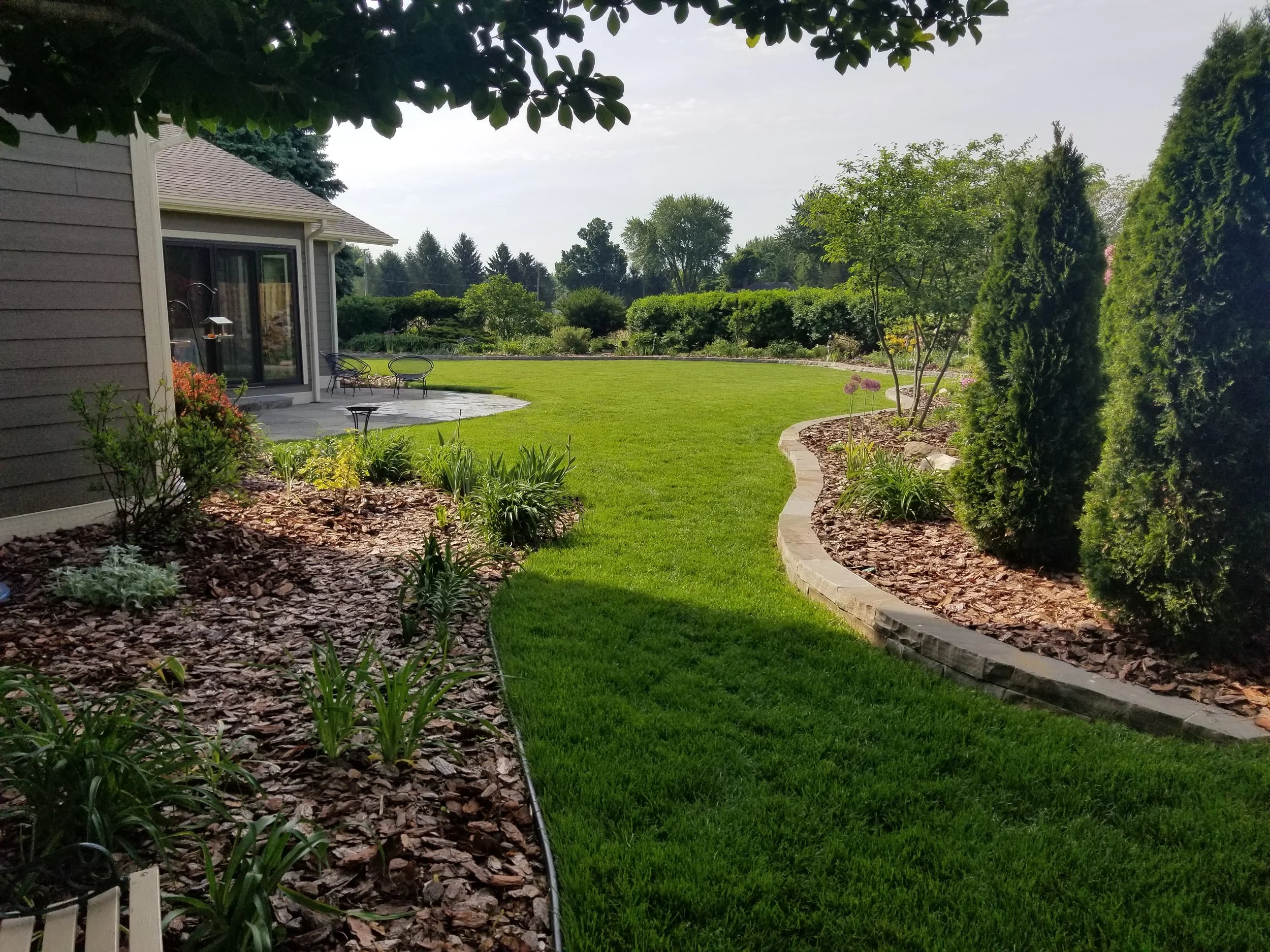 View of a landscaped backyard with a green lawn, flower beds edged with bricks, and trees, with a house and patio furniture in the background.