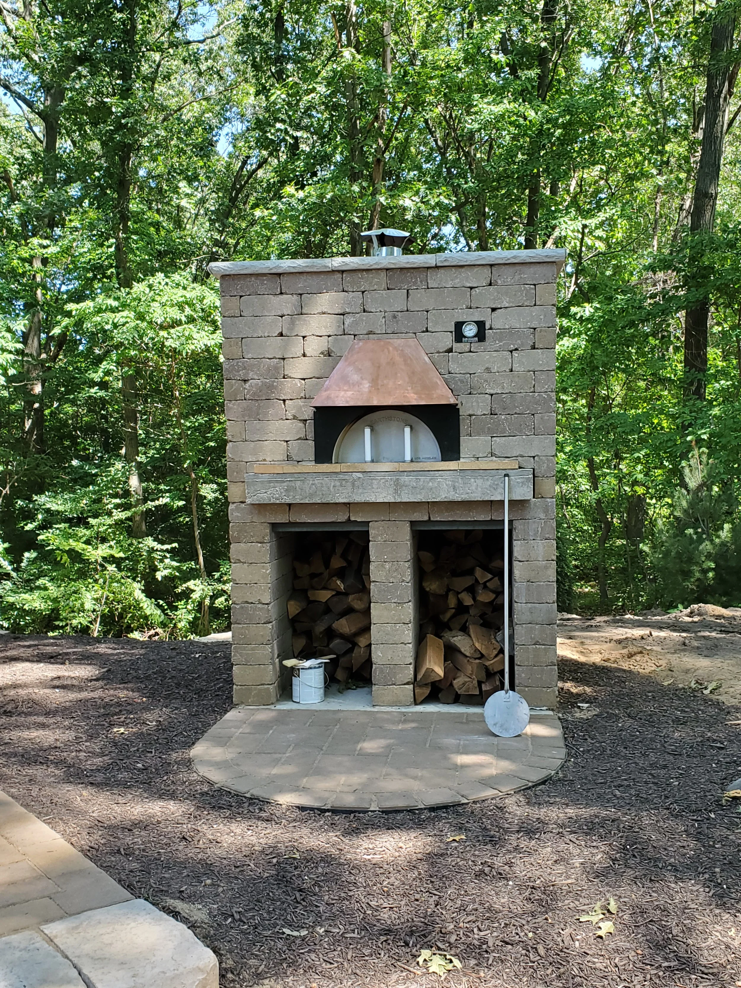 An outdoor brick oven with a chimney, stacked firewood underneath, and a cooking surface. A metal pizza peel leans against the right side of the oven, and a small bucket with a spatula is placed on the ground nearby. The area is surrounded by trees a