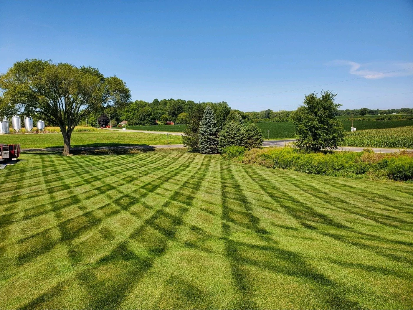 A well-maintained, striped green lawn with shadows from nearby trees, surrounded by trees and fields under a clear blue sky.