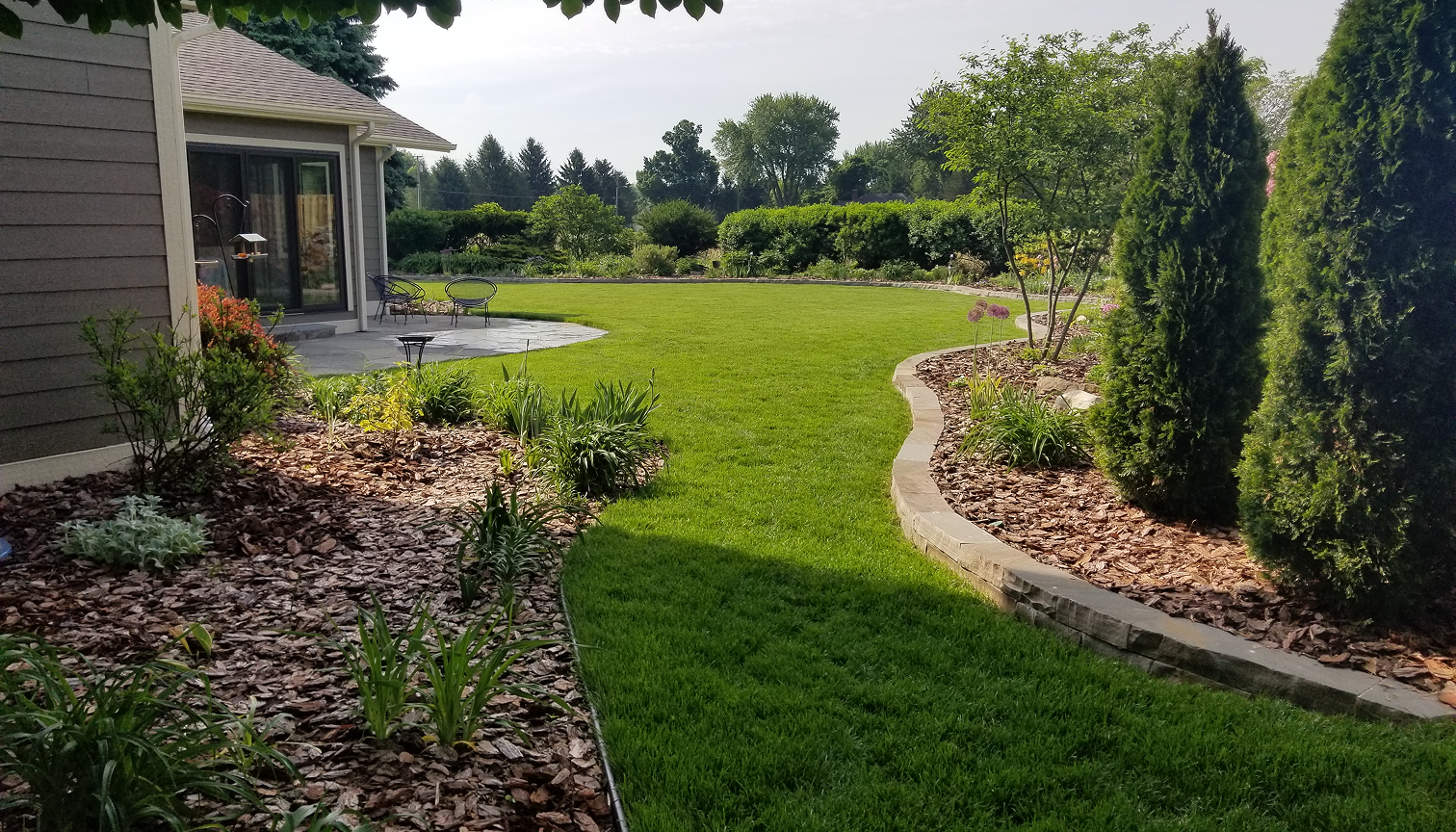 A well-maintained backyard with a green lawn, landscaped flower beds, trees, shrubs, and a patio with outdoor furniture next to a house.