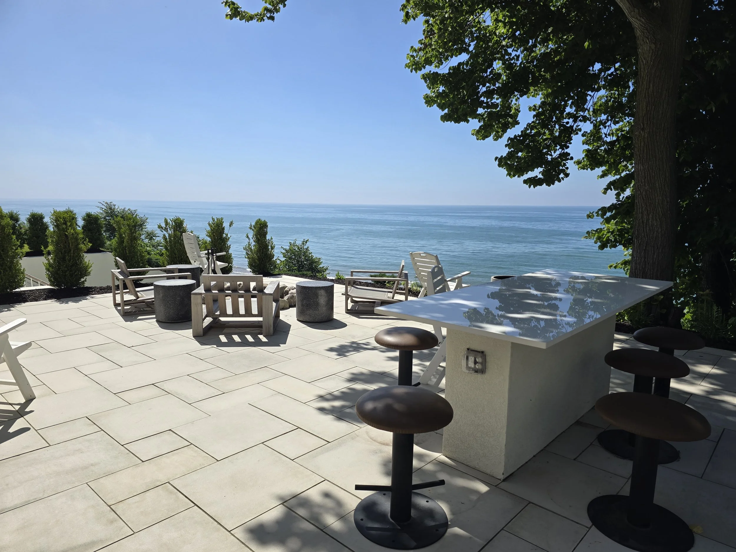 Outdoor patio overlooking the ocean with white chairs, a table, and bar stools under shade of a large tree.