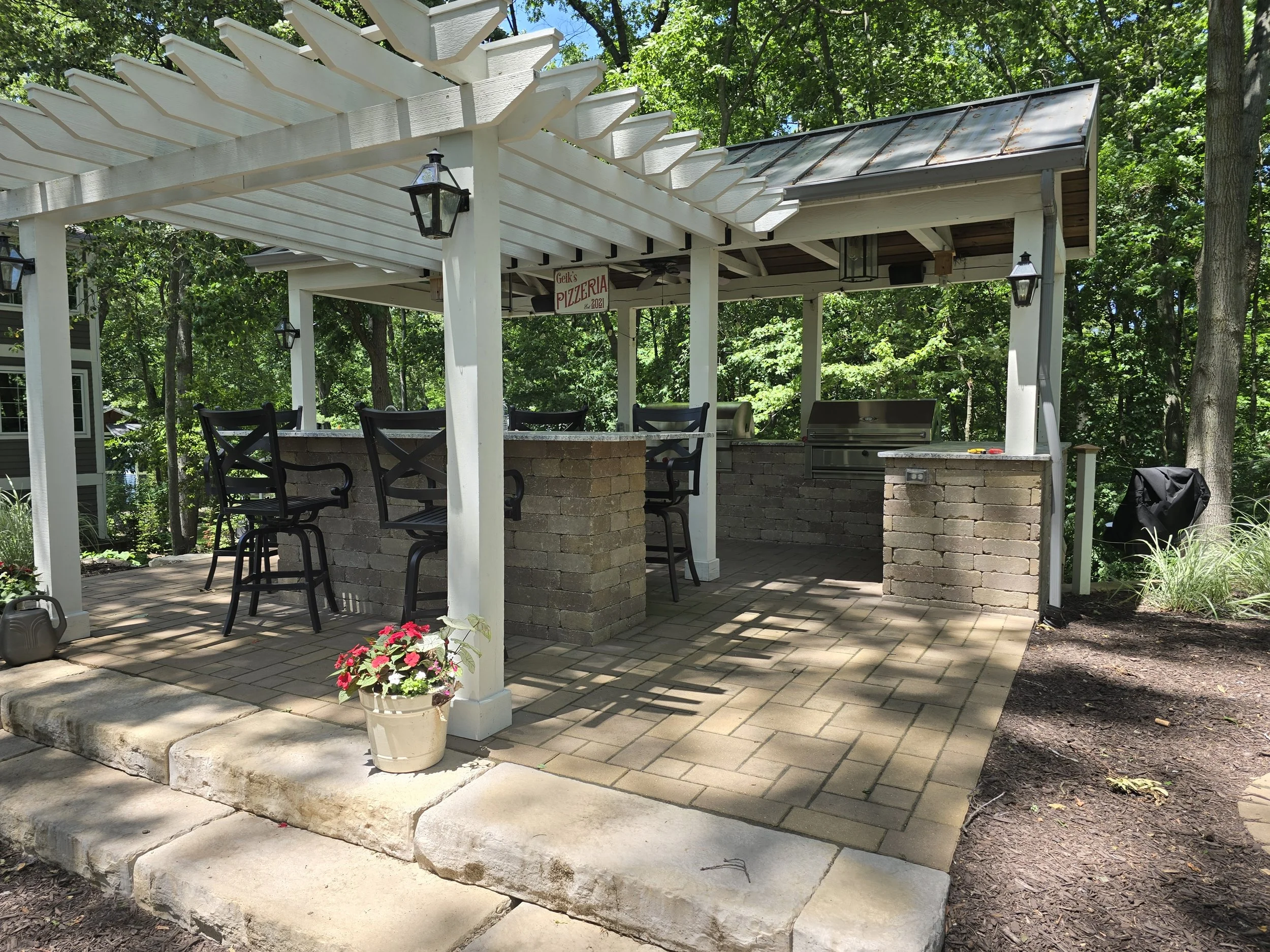 An outdoor patio area with a white pergola, bar counter, barstools, and a grill, surrounded by green trees.