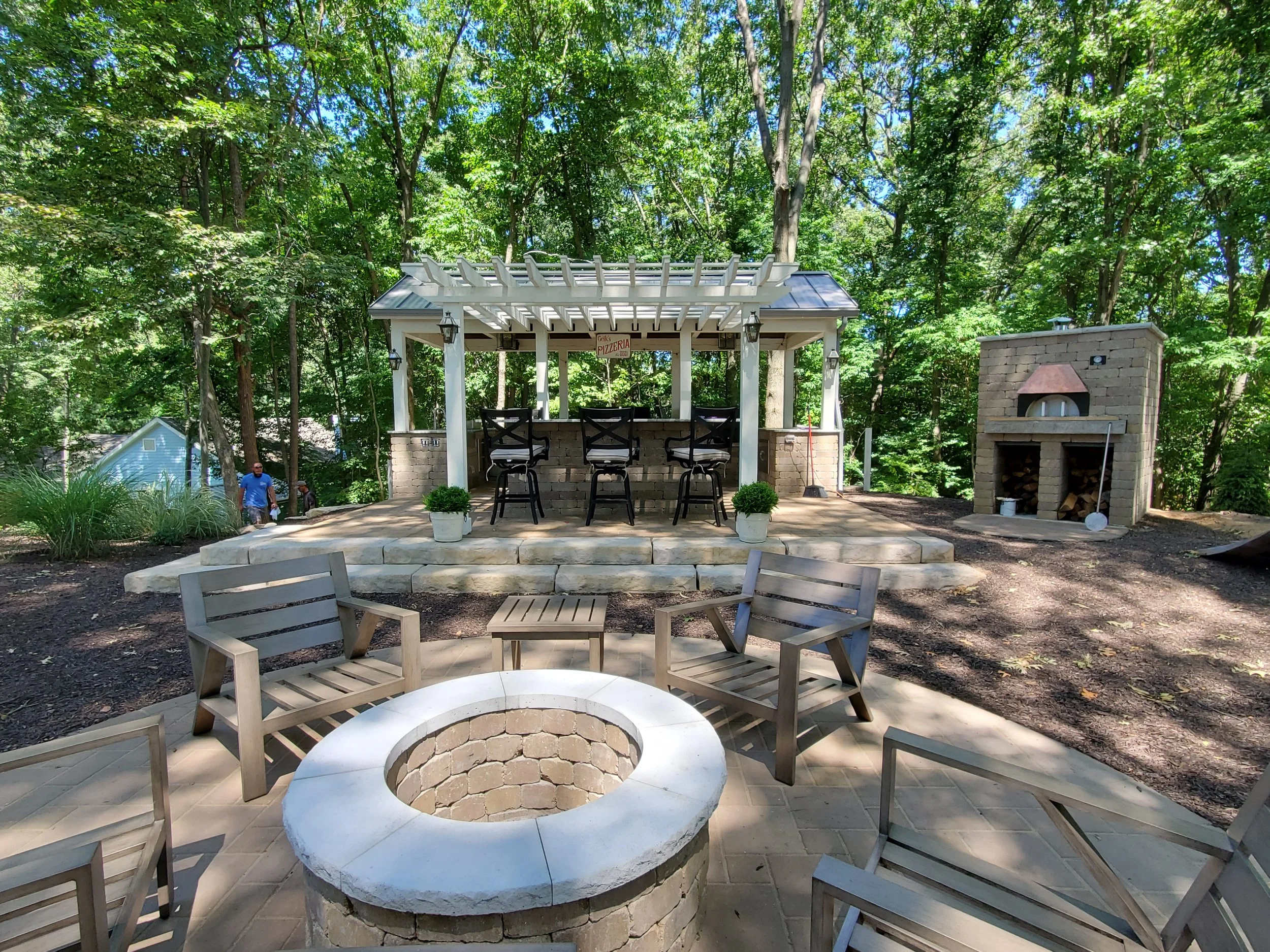 Outdoor backyard seating area with a fire pit in the foreground, a raised patio with bar stools beneath a pergola in the middle, and a stone pizza oven on the right, all surrounded by trees.