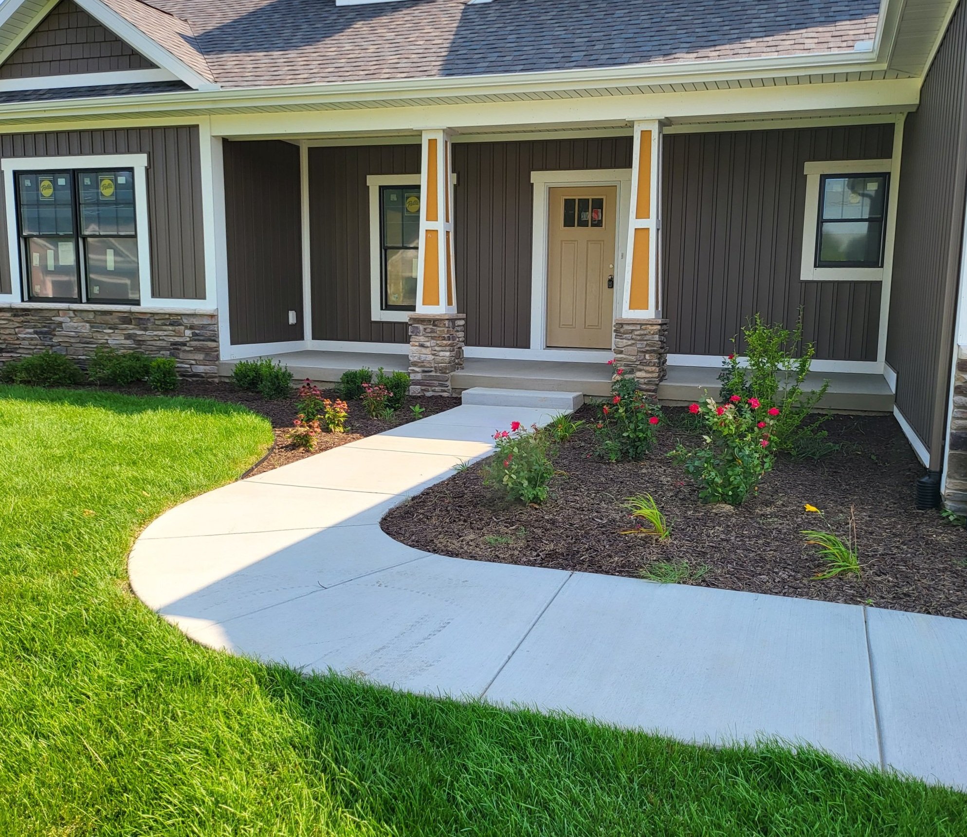Front porch of a house with a curved concrete walkway, green lawn, and planted flower bed with pink flowers.
