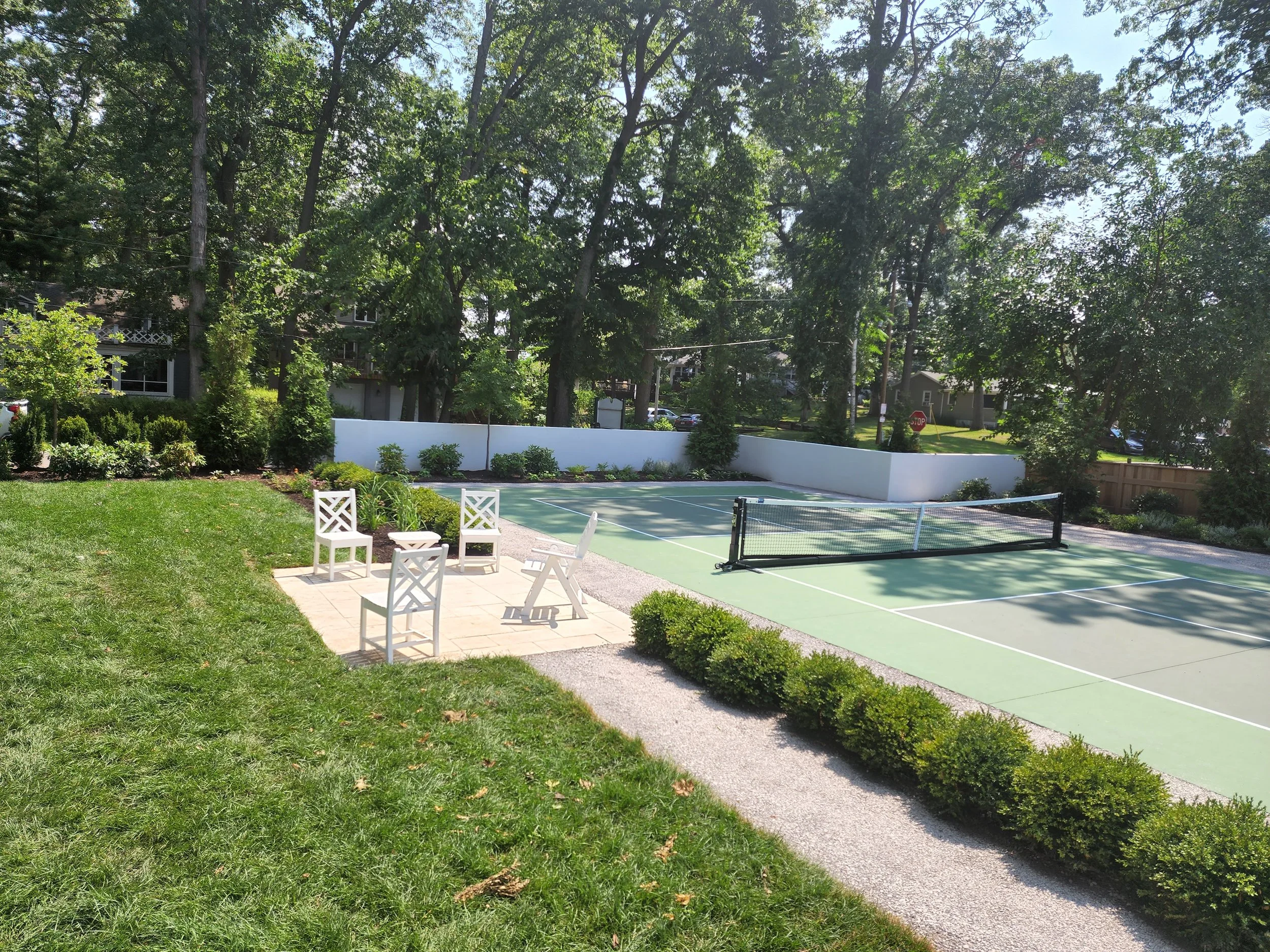 A backyard with a tennis court surrounded by trees and greenery, with a small patio area featuring four white chairs and a small round table.