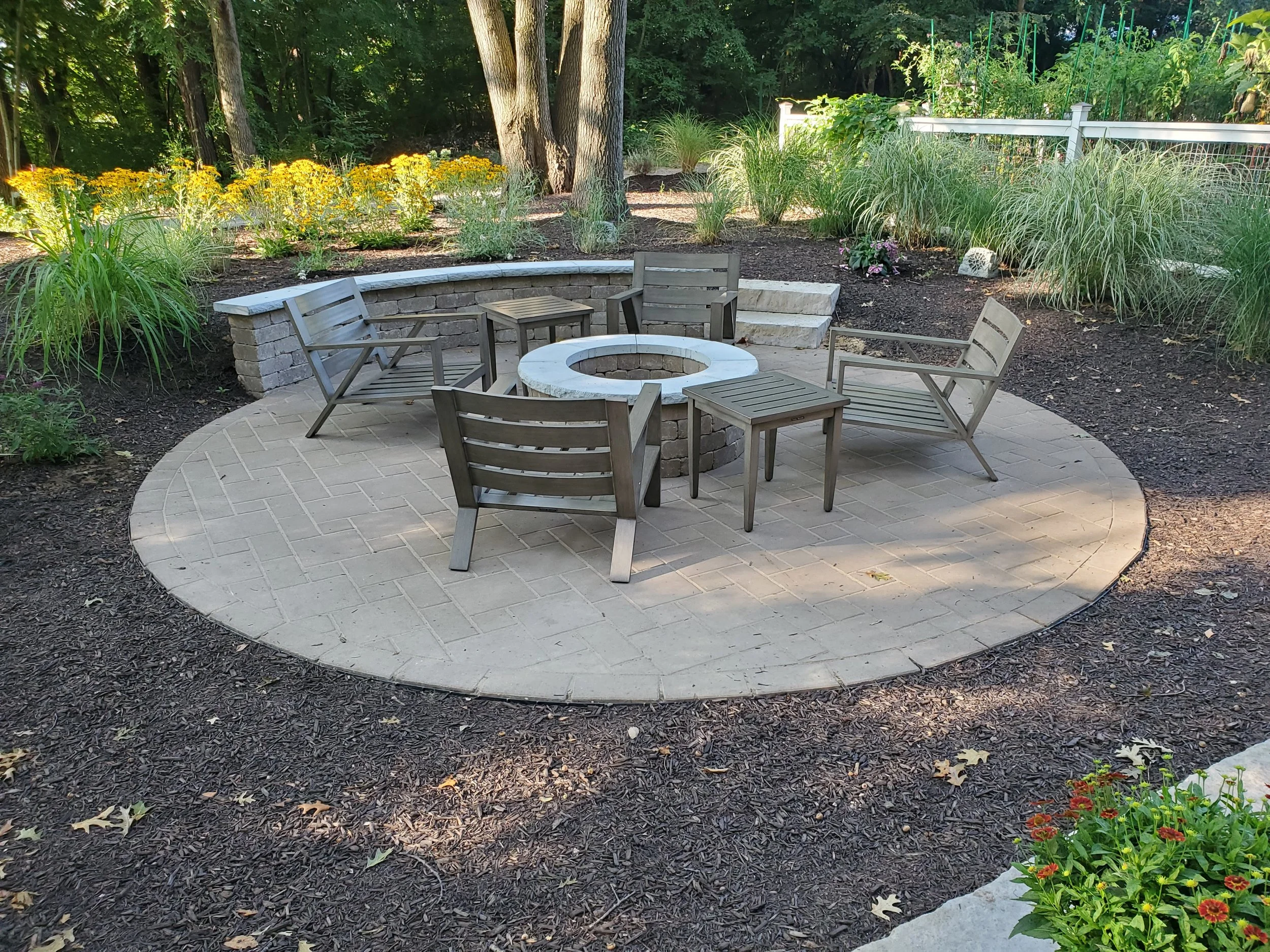 Outdoor fire pit area with four wooden chairs, small tables, and landscaped garden with flowers and trees.