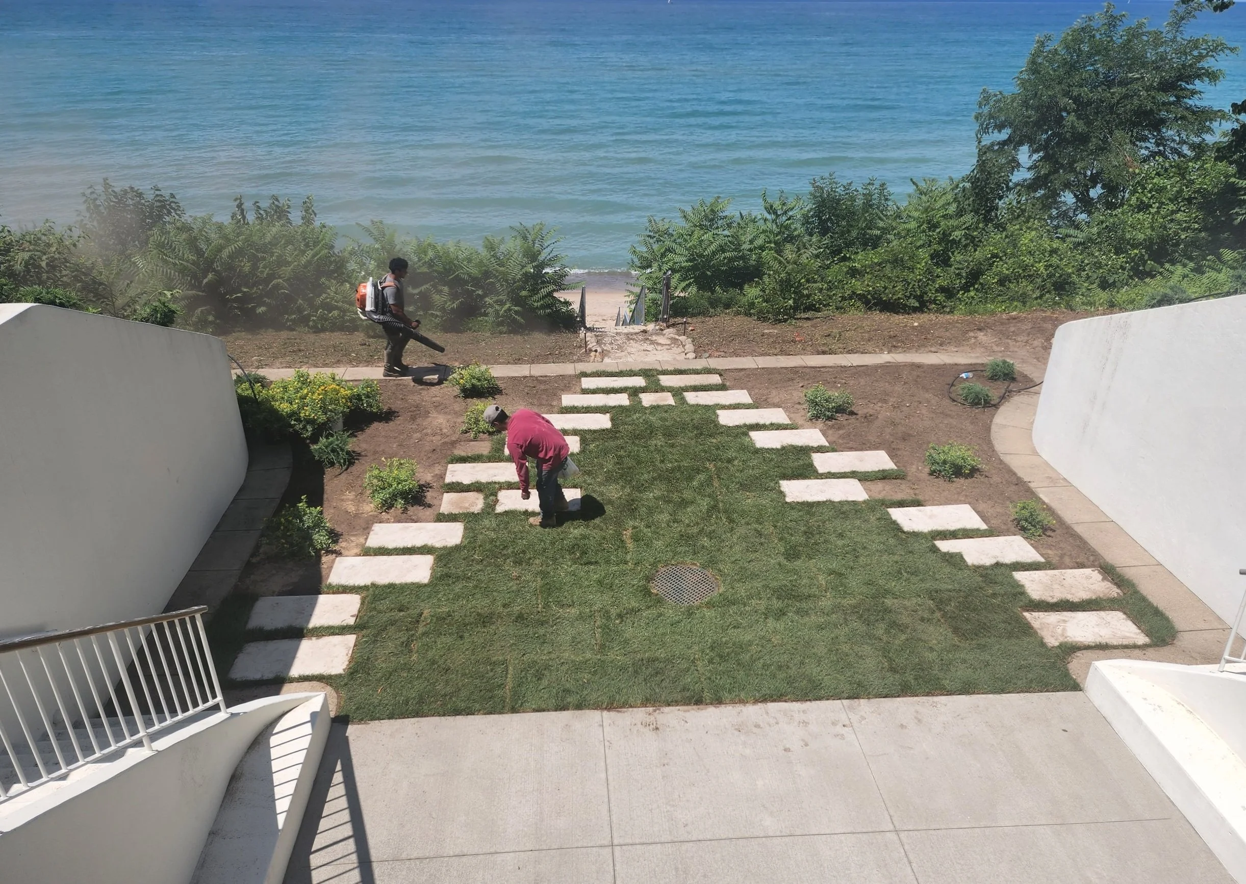 Two people planting grass and landscaping near a beach, with stairs leading to the sand and ocean, surrounded by bushes and white walls.