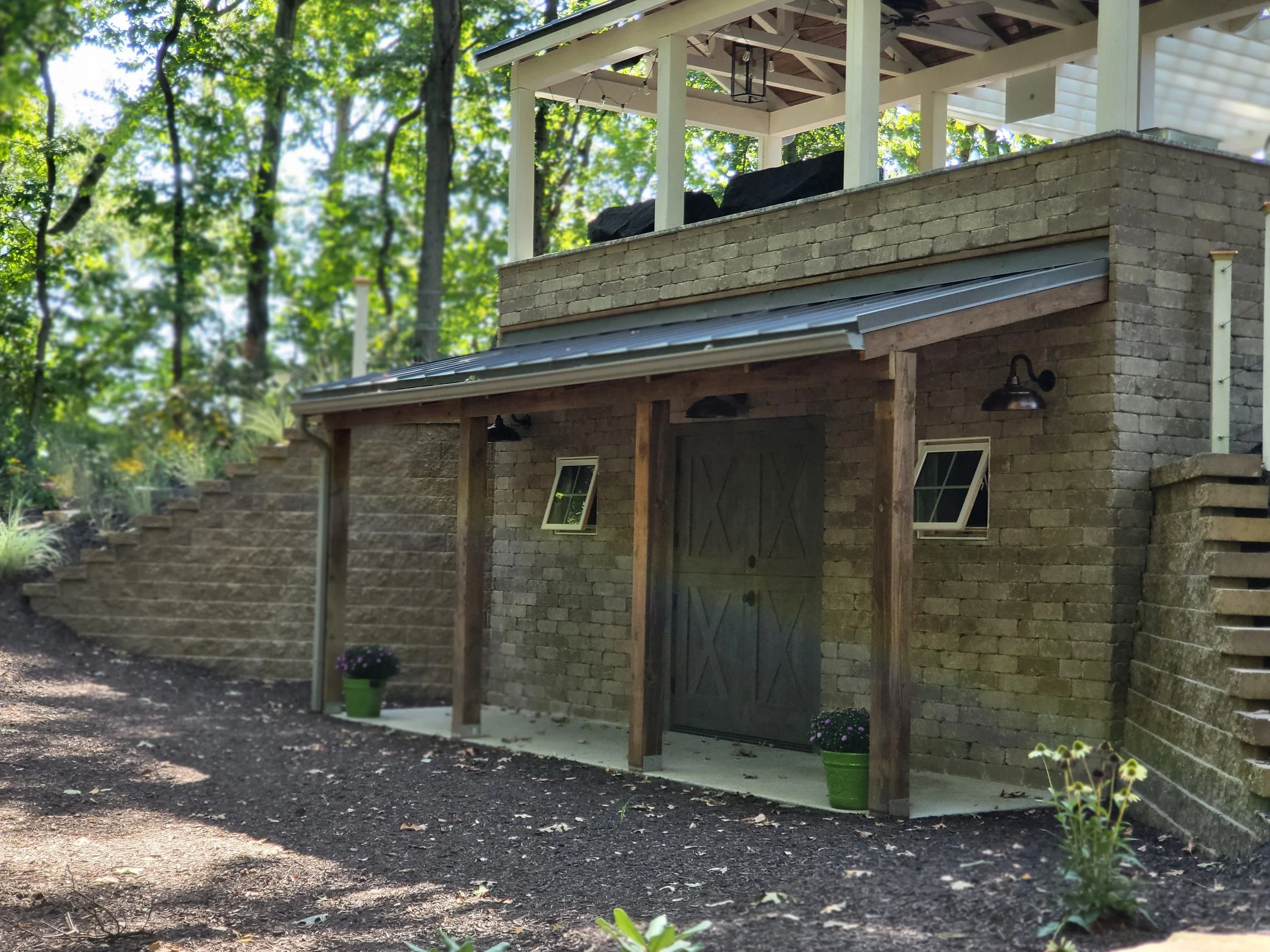 A brick building with a wooden door, two small windows, and outdoor wall lights, surrounded by trees and plants.