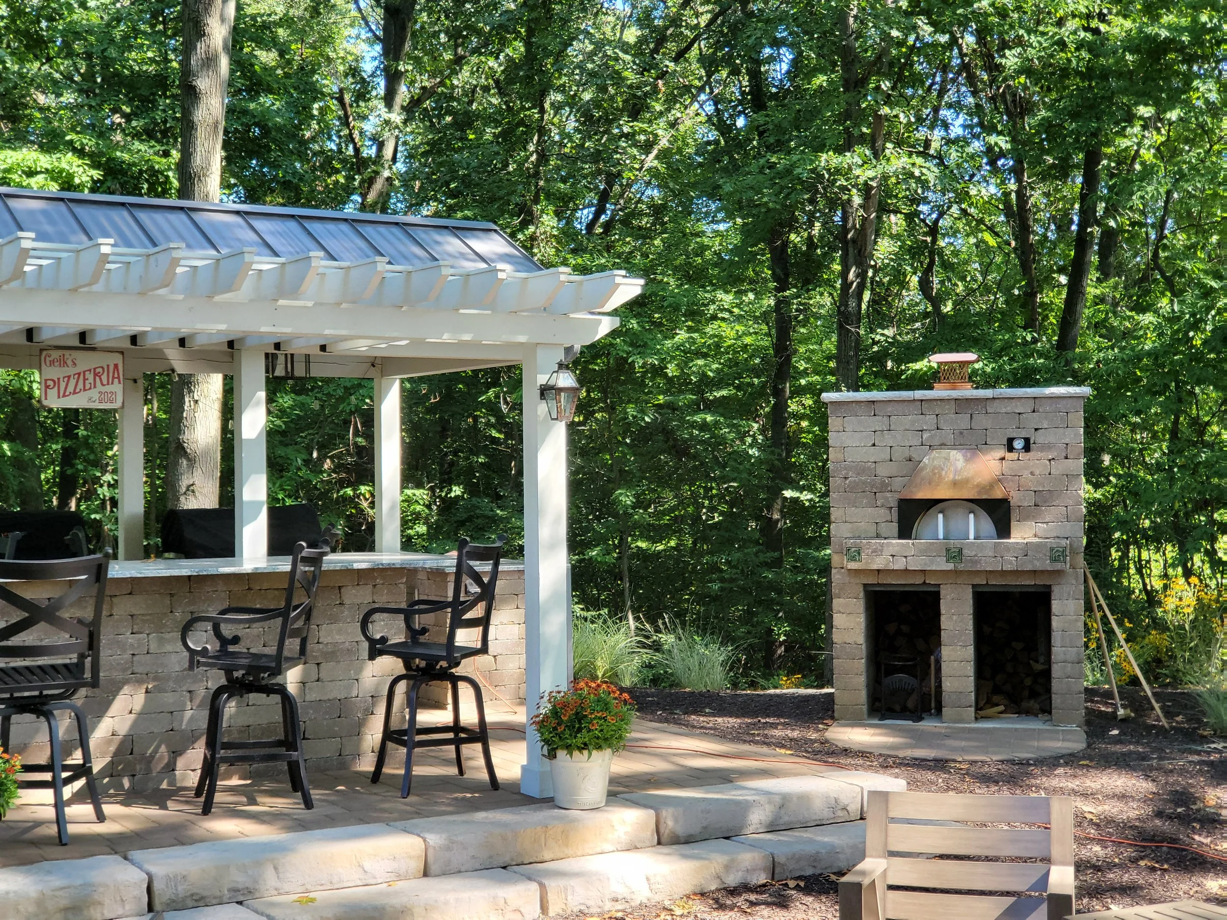 Outdoor patio area with a brick pizza oven, a covered bar with barstools, potted flowers, and lush green trees in the background.
