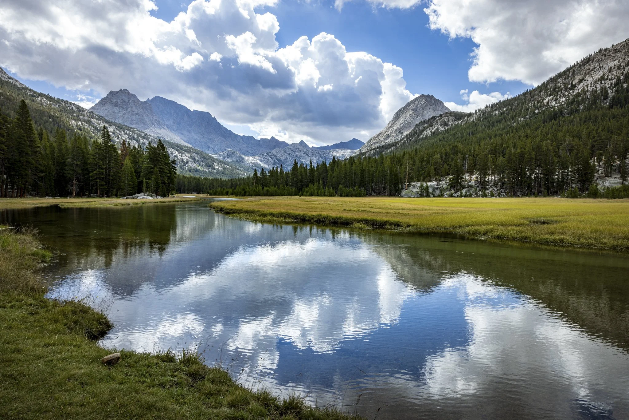 Scenic view of a river reflecting clouds and trees, surrounded by mountains and dense forest under a partly cloudy sky.