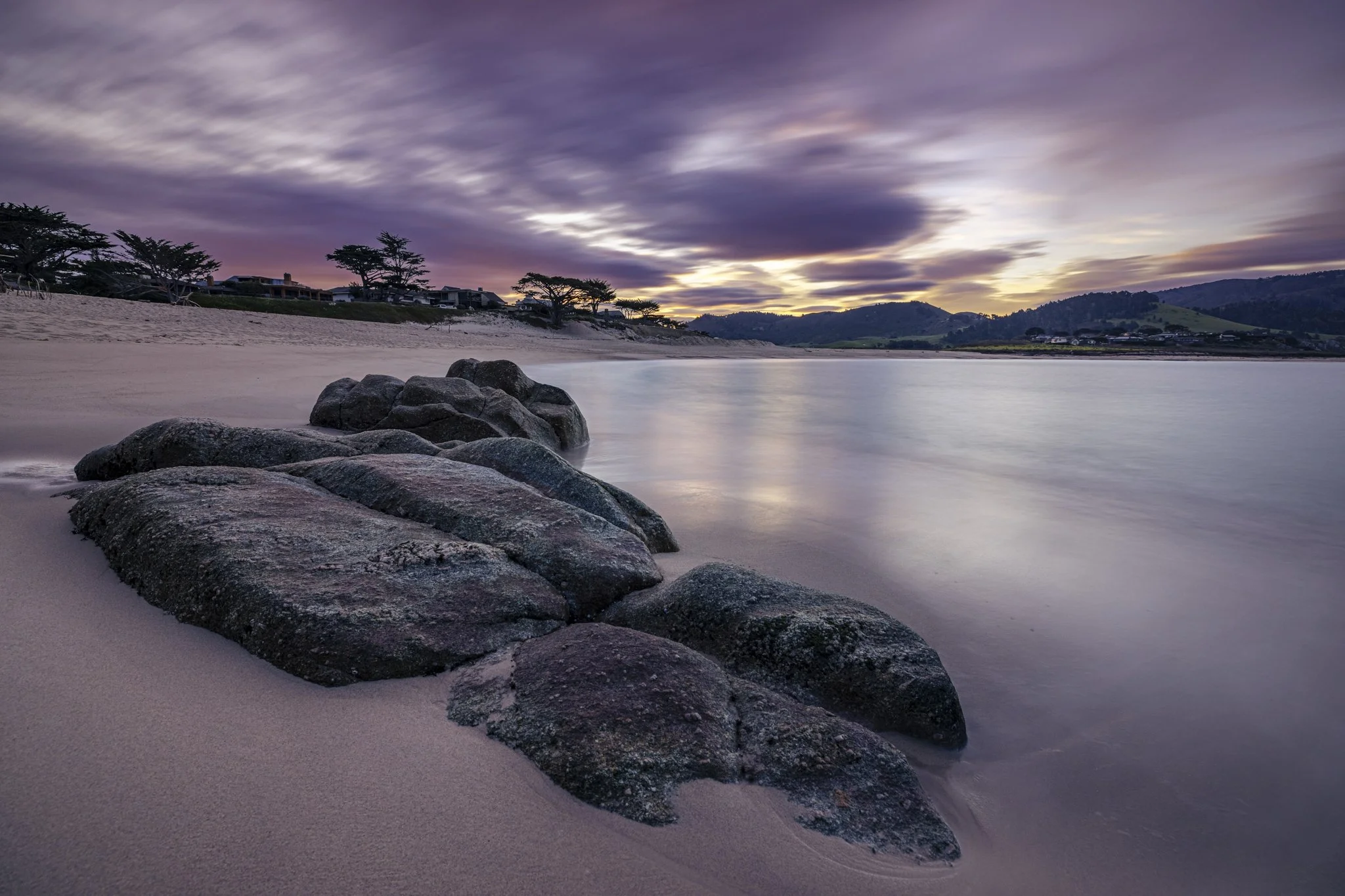 A peaceful beach scene at sunset with purple clouds, calm water, rocks on the sandy shore, and trees in the distance.