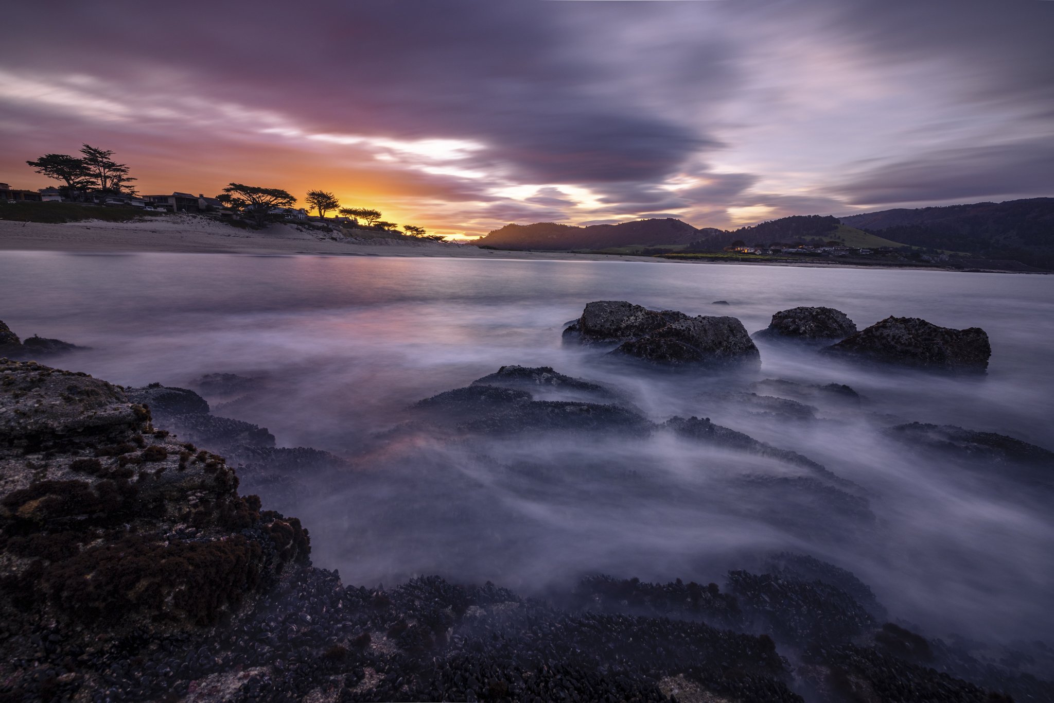 A long exposure of a beach at sunset with rocks in the foreground, water flowing over the rocks, and trees and hills in the background under a partly cloudy sky.