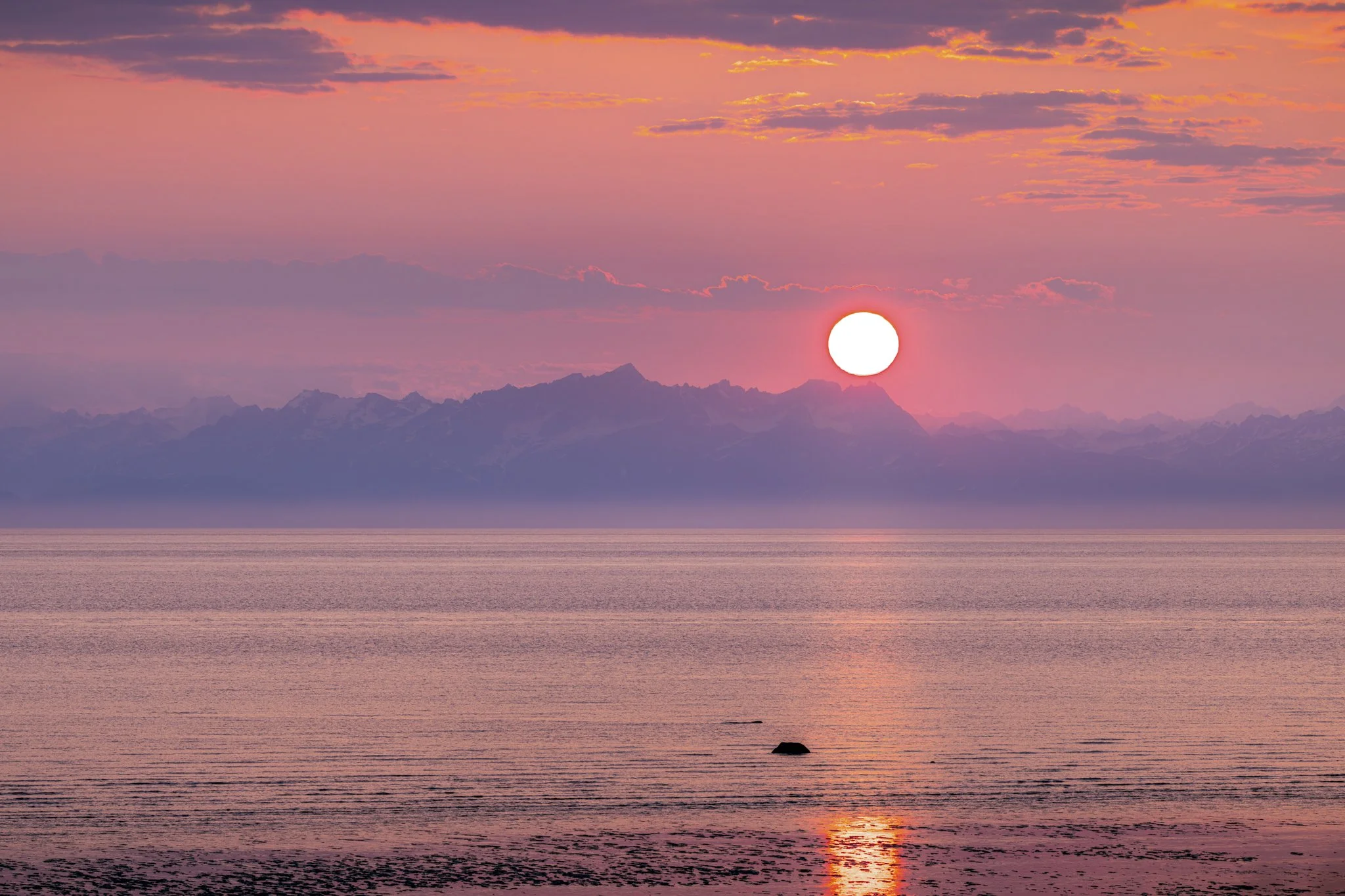 Sunset over a calm body of water with mountains in the background and pink, purple, and orange sky.