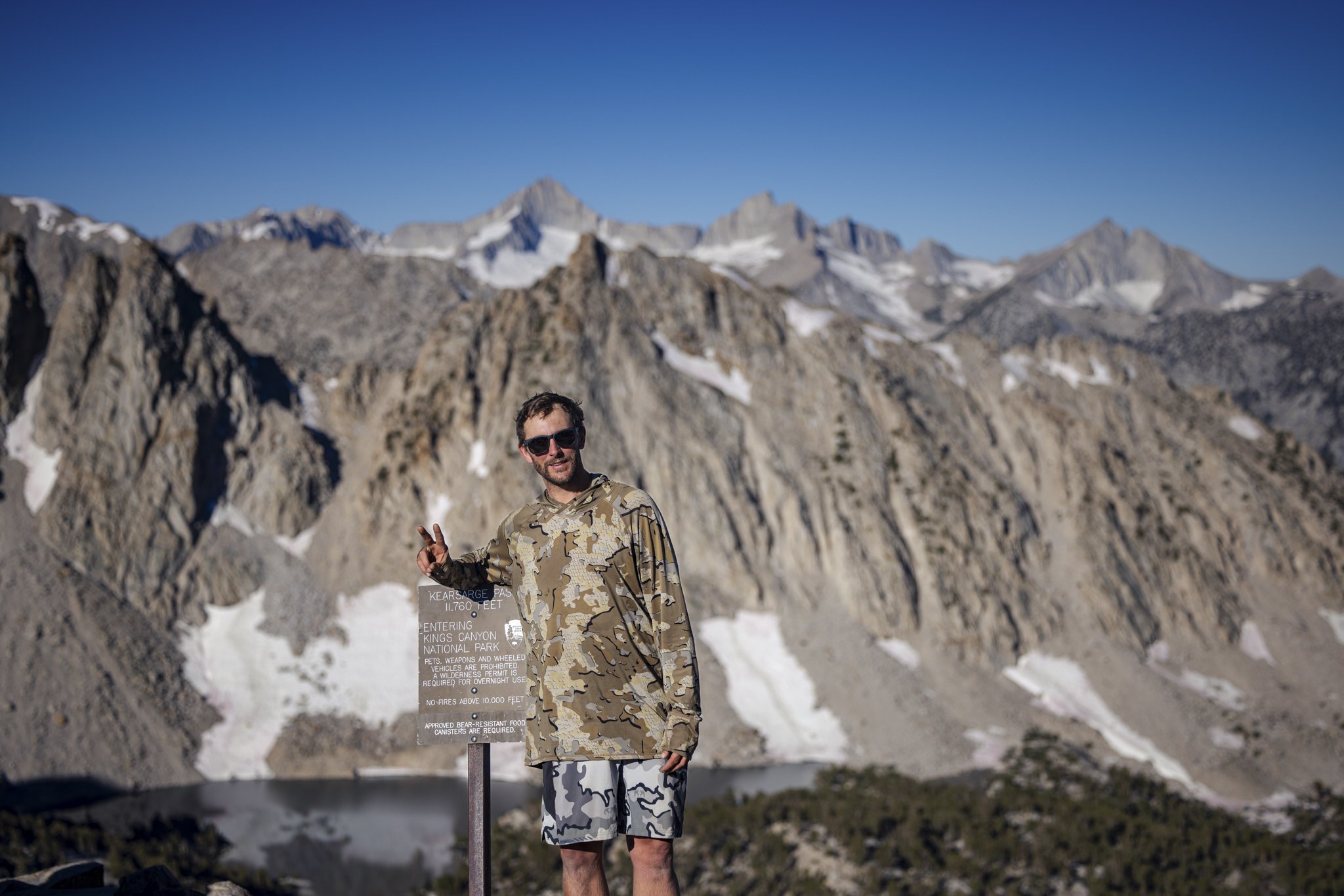 A man making a peace sign while standing next to a sign at Kings Canyon National Park with rugged mountains and patches of snow in the background.