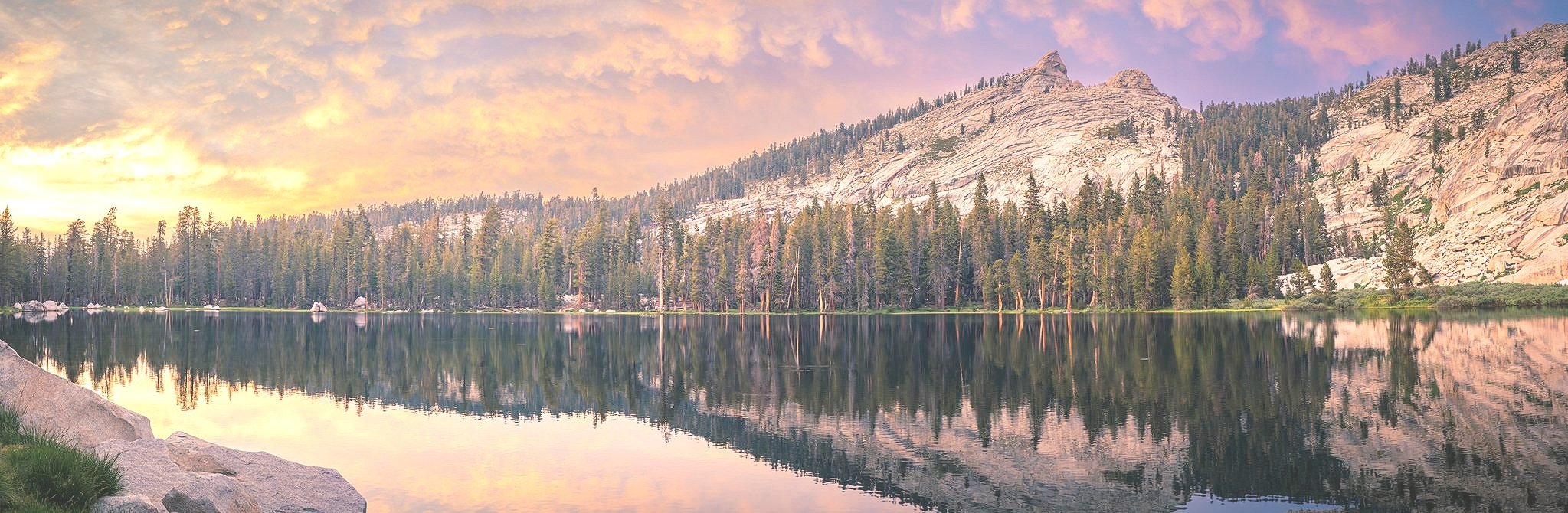A serene mountain lake with a calm reflective surface, surrounded by a dense pine forest, with rocky cliffs in the background and a colorful sky at sunset or sunrise.