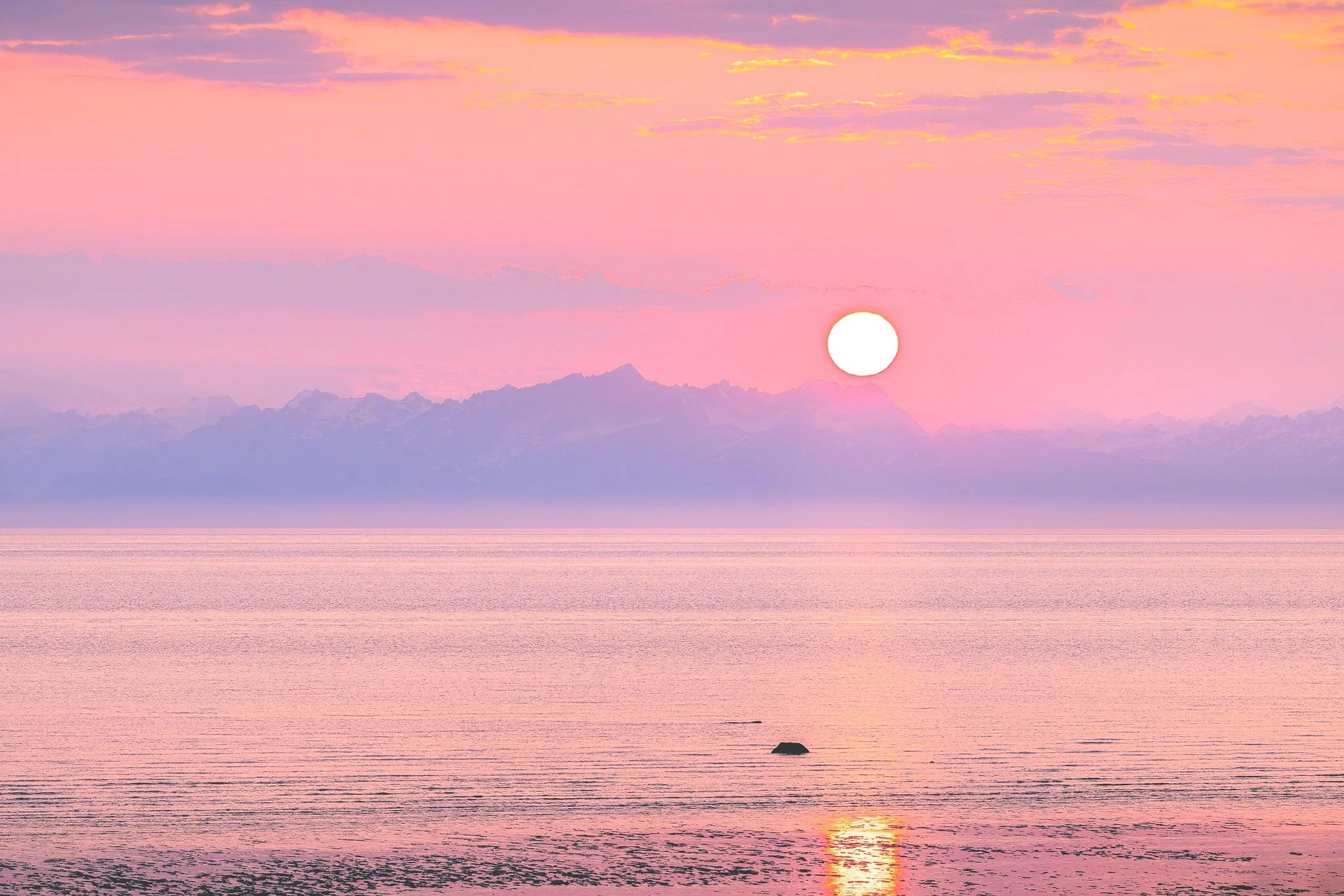 Sunset over pink and purple water with mountains in the background and rocks in the water