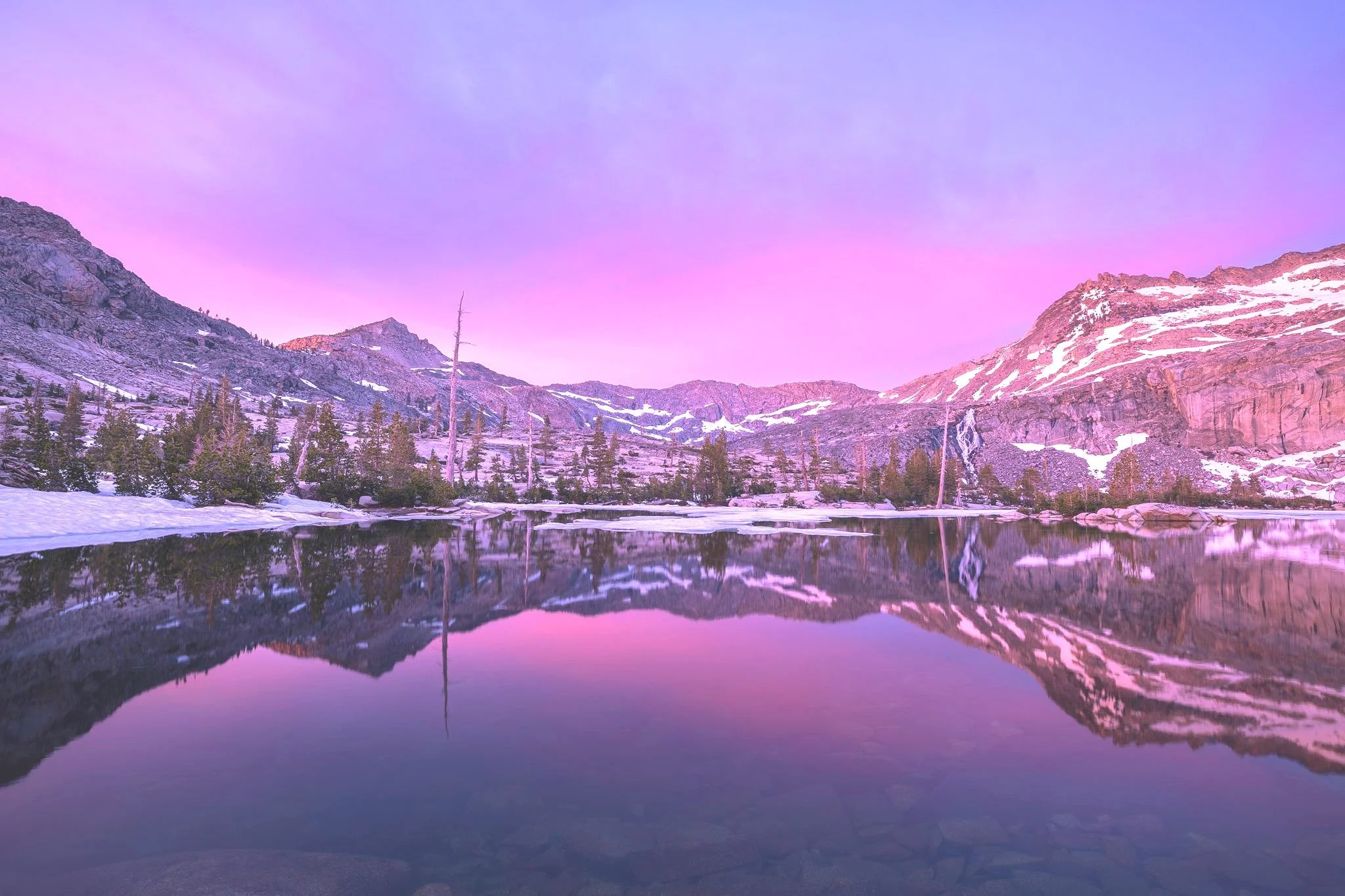 A serene snowy mountain landscape reflecting in a calm lake under a colorful pink and purple sunset sky.