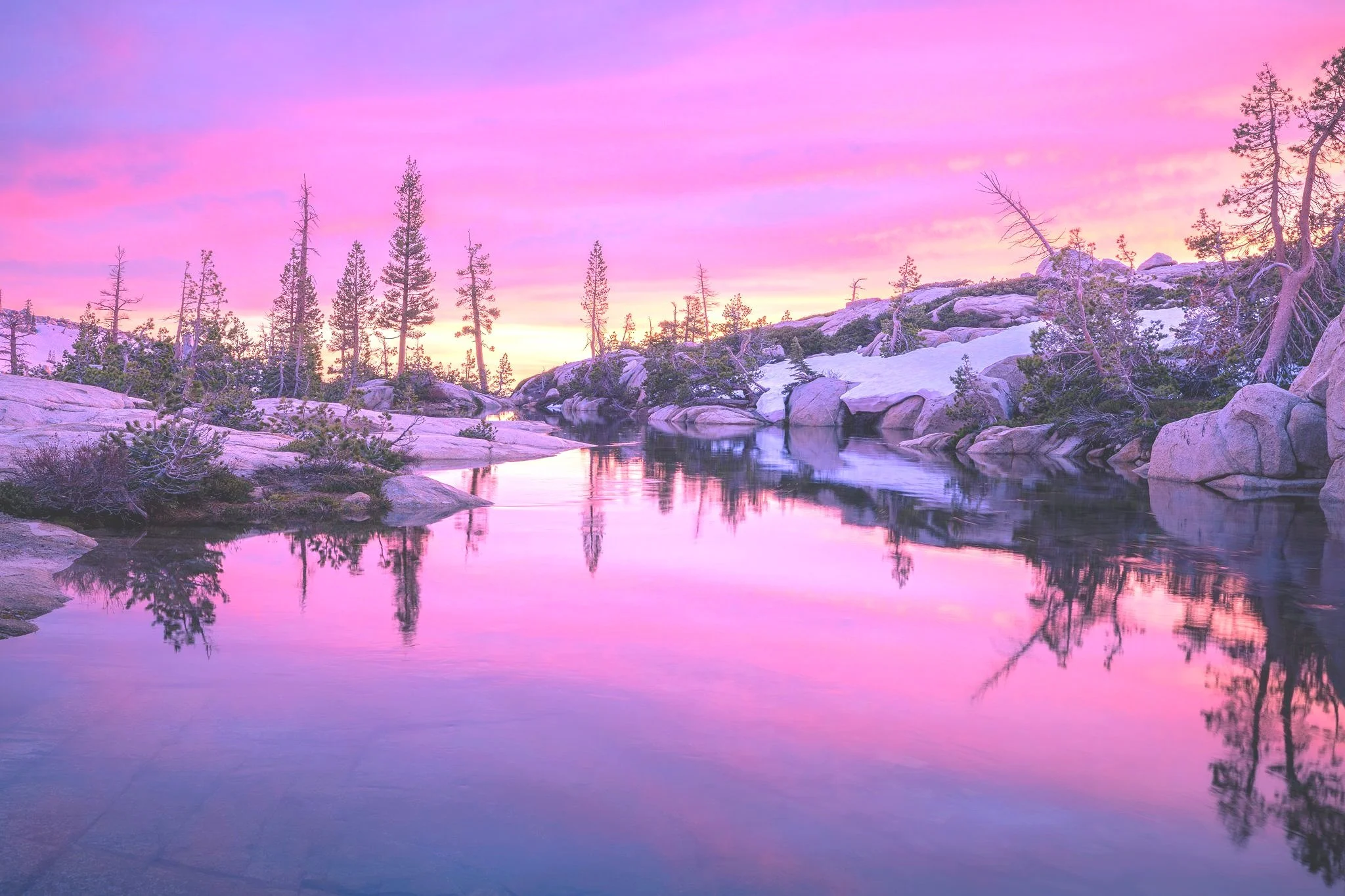 A serene mountain landscape with a calm river reflecting the colorful pink and purple sky, surrounded by snow-covered rocks and tall pine trees.