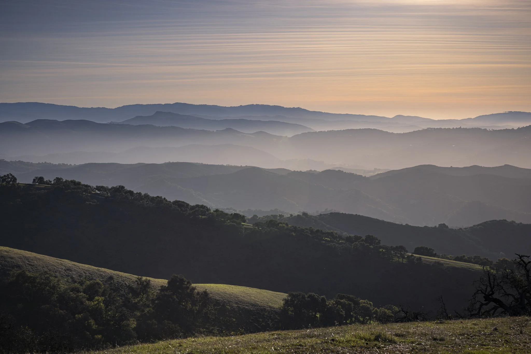 Multiple layers of rolling hills and mountains with a hazy atmosphere, under a sky with soft pink and purple hues.