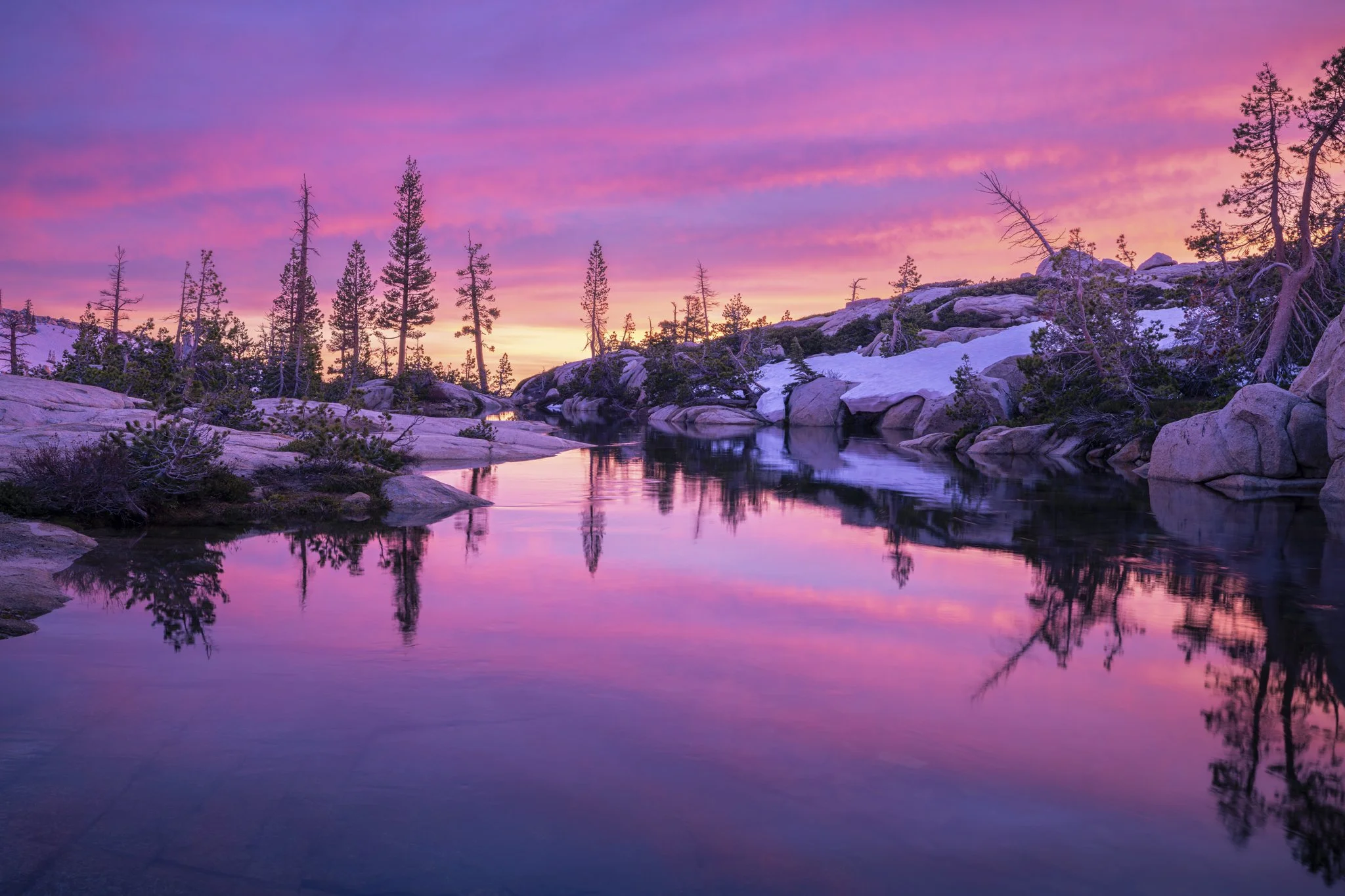 A serene landscape of a mountain lake at sunset, with snow-capped rocks and pine trees along the shoreline, and a sky painted in shades of pink and purple reflected in the calm water.