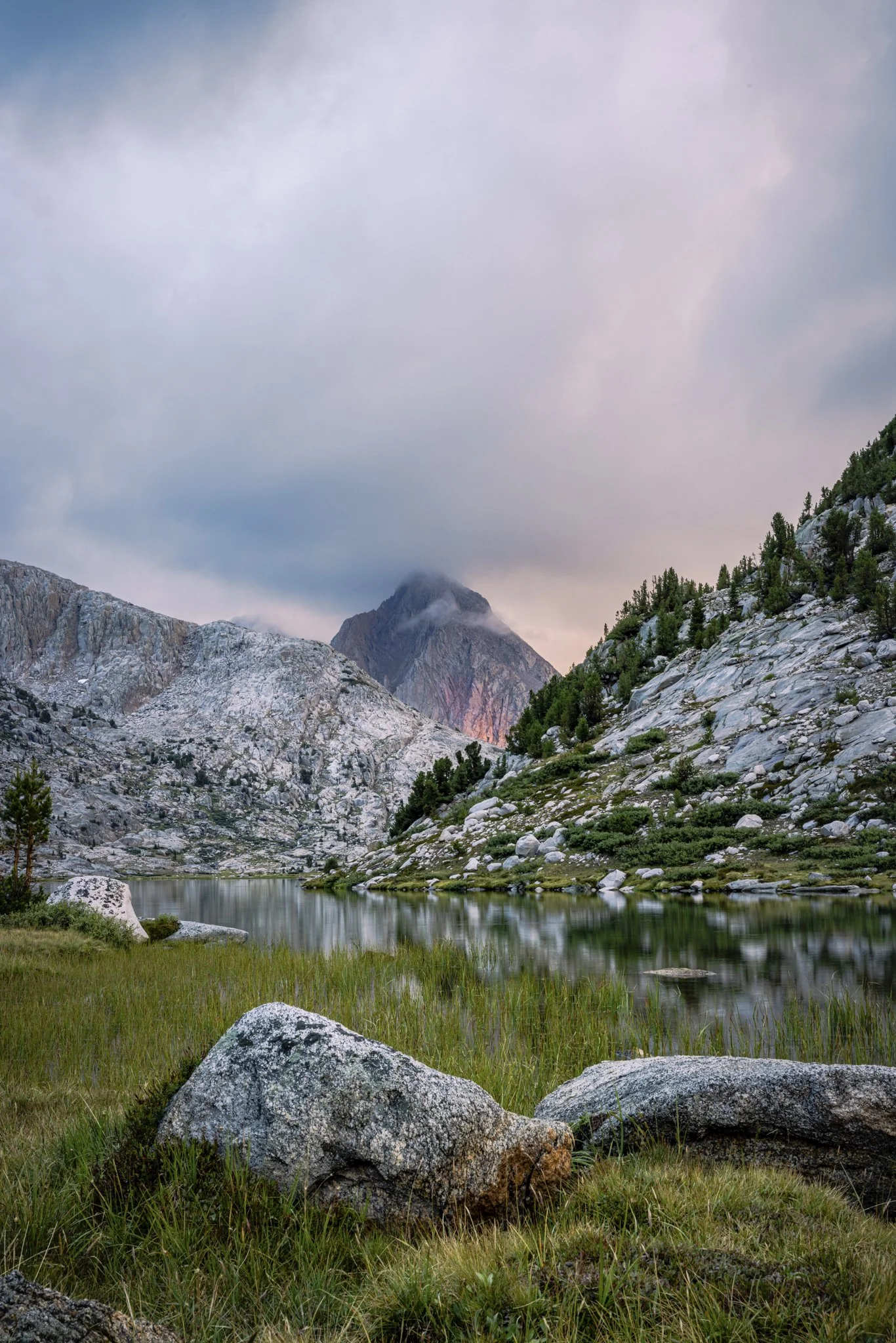 Scenic mountain landscape with rocky terrain, green grass, trees, a calm lake, and a cloudy sky.