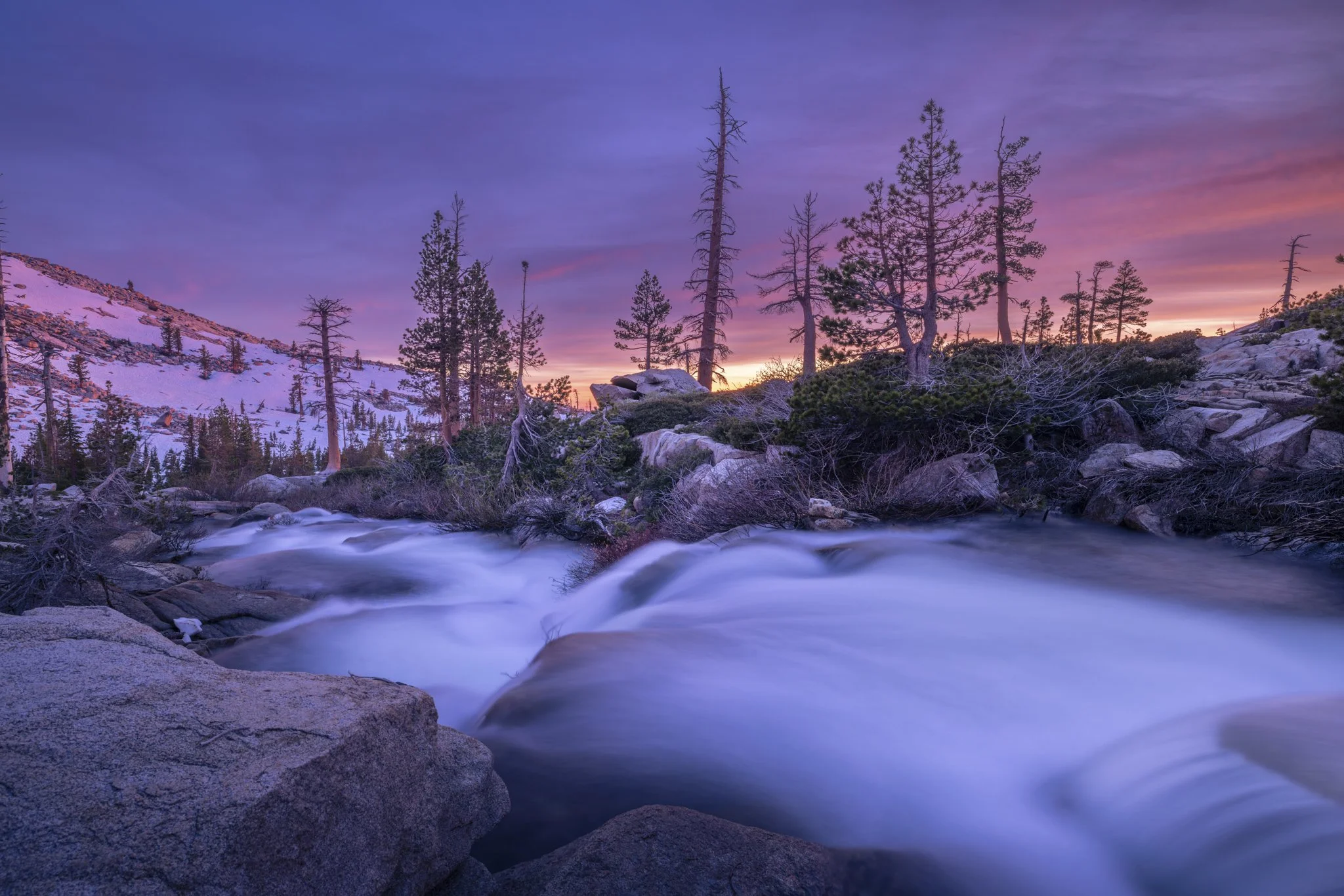 A flowing mountain river at sunset with snow on distant hills and tall pine trees under a colorful purple and pink sky.