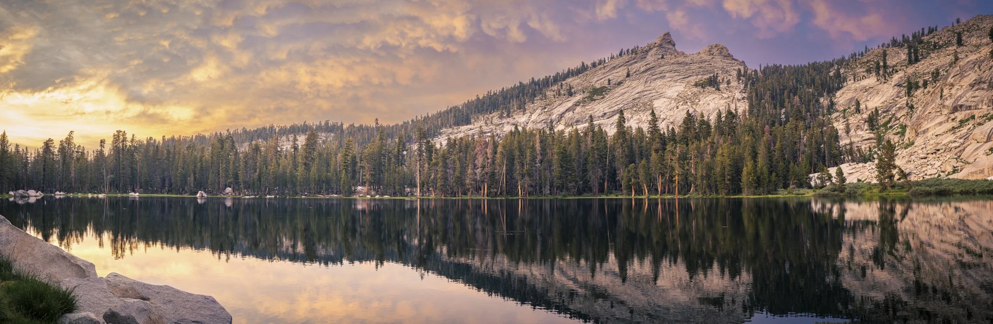 A serene mountain landscape at sunset with a still lake reflecting the sky, surrounded by dense pine trees and rocky mountain slopes.
