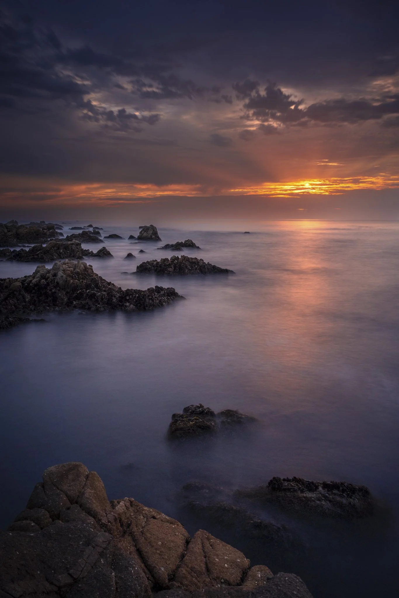 Sunset over the ocean with rocks in the foreground and purple-blue sky with clouds.