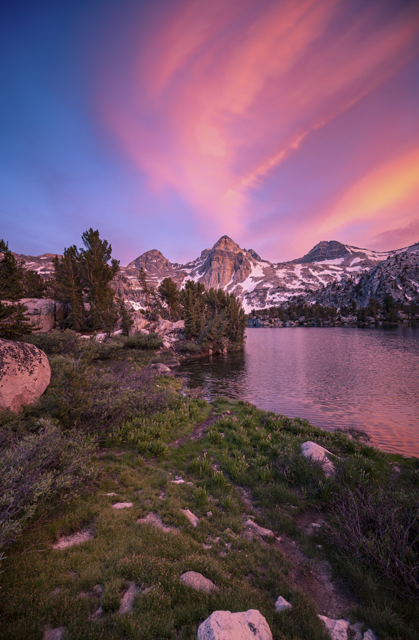 A tranquil mountain scene during sunset with snow-capped peaks reflecting pink and purple hues, green pine trees along a lake shoreline, and a grassy trail in the foreground.