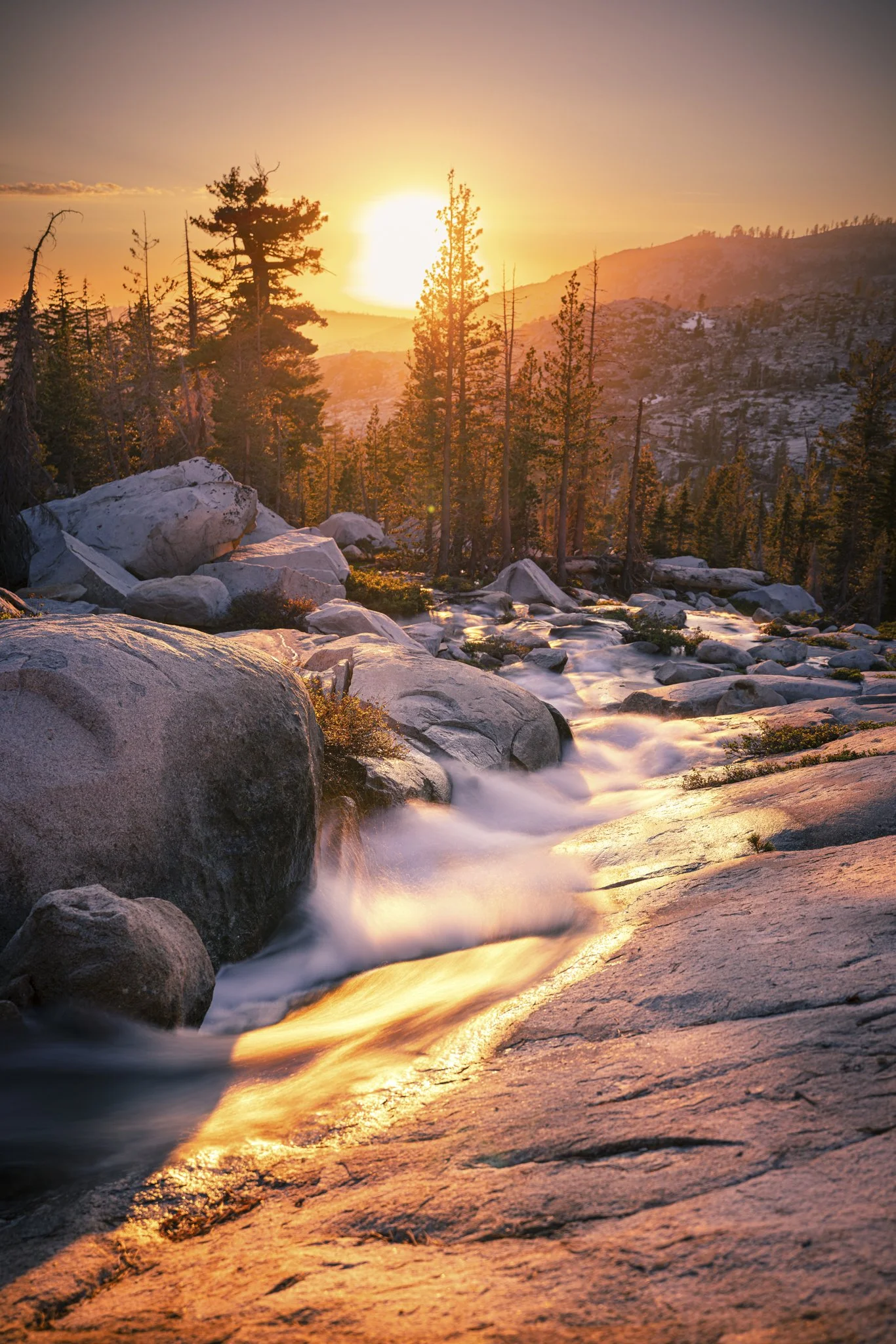 A scenic sunset over a mountain landscape with a flowing creek, pine trees, rocks, and a glowing sky.