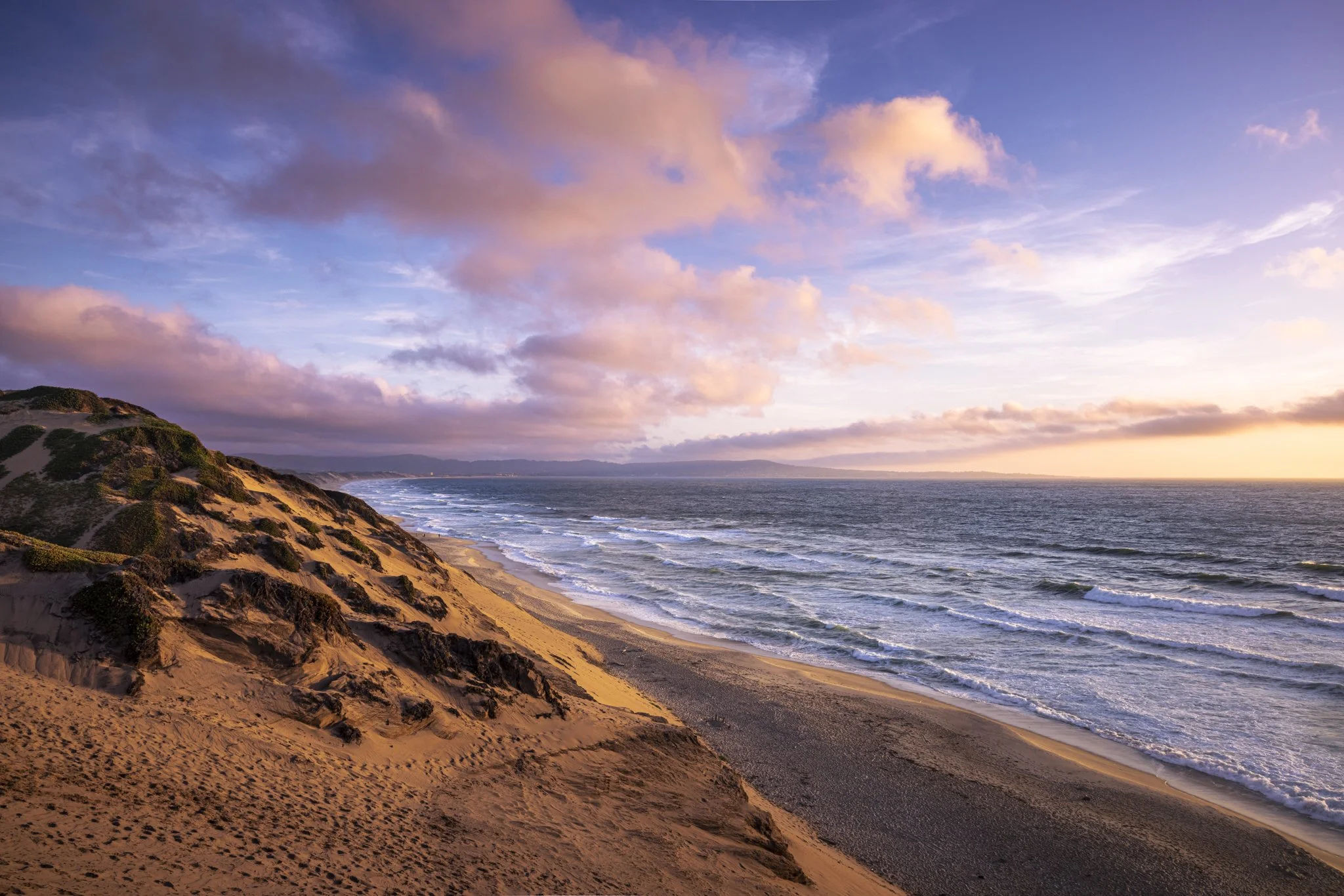 Sunset over a sandy beach with waves crashing and a sky filled with pink and purple clouds.