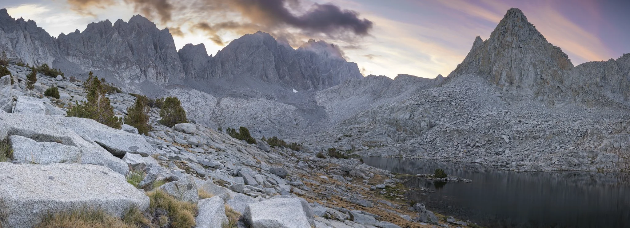 Scenic mountain landscape at dusk with rocky slopes, jagged peaks, a small lake, and colorful sky with clouds.