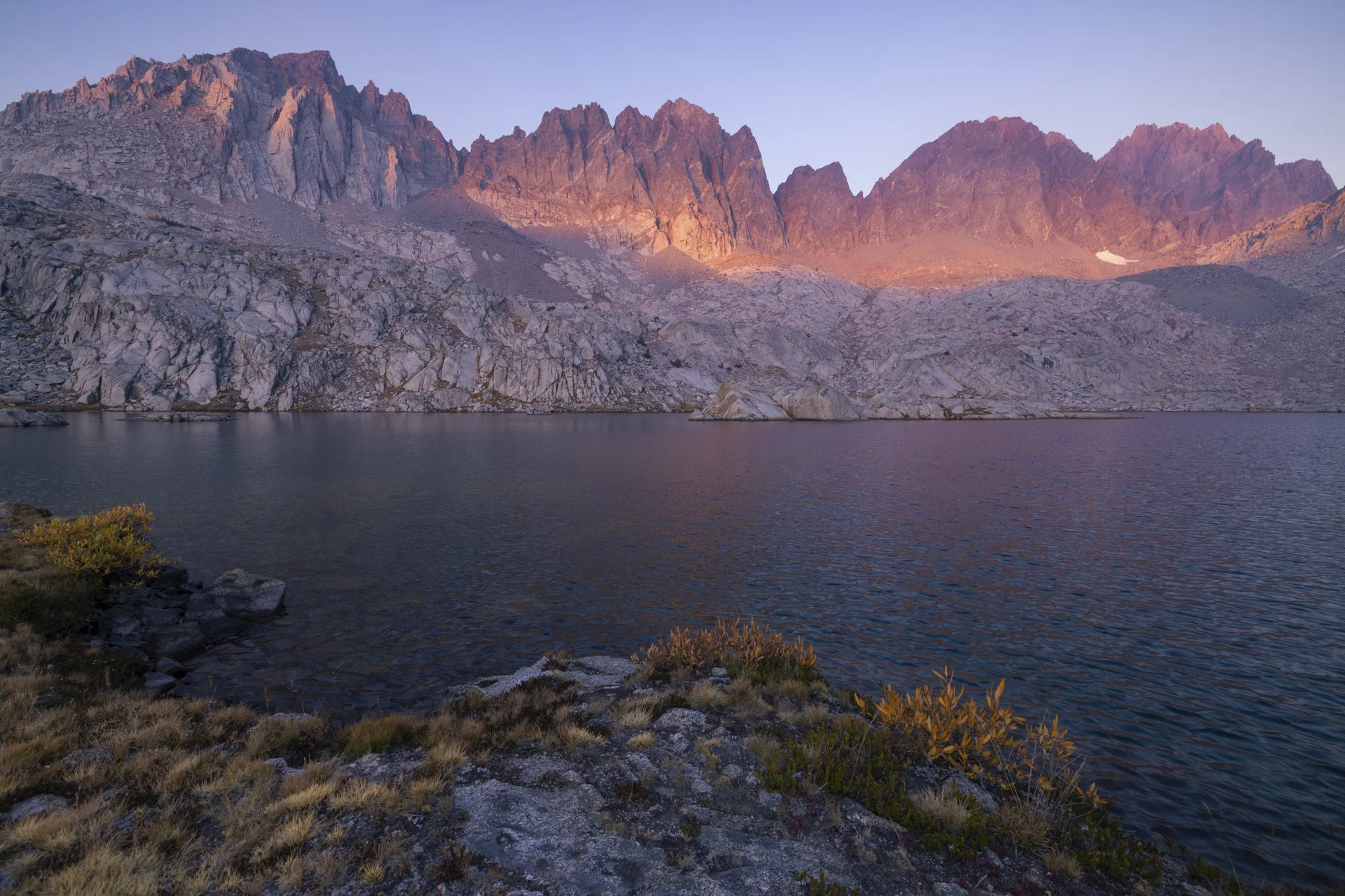 Mountain lake scene at dusk with rocky shore, calm water, and rugged mountain peaks in background