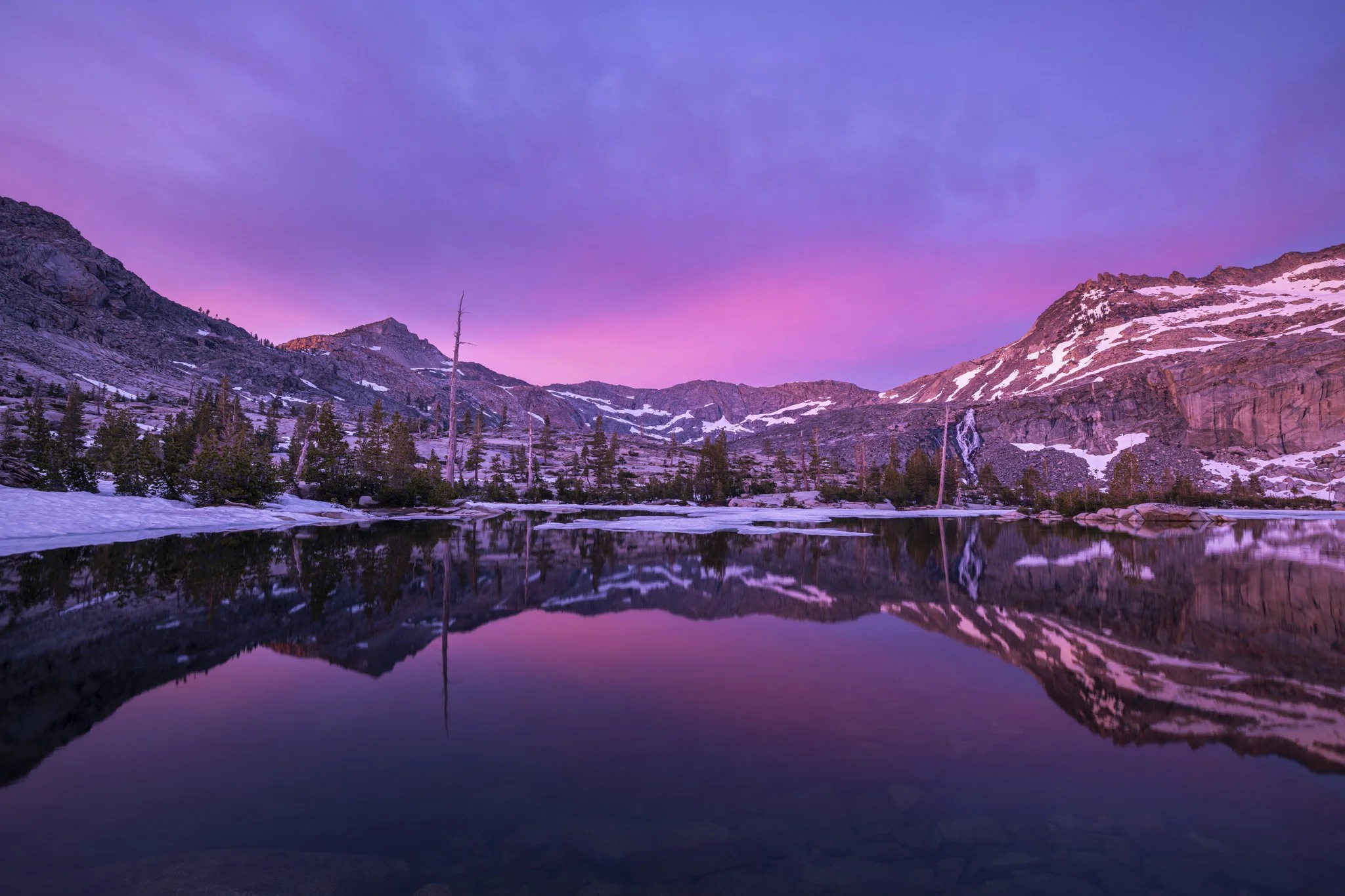 Snow-covered mountain landscape with a lake reflecting the colorful purple and pink sky at dusk.