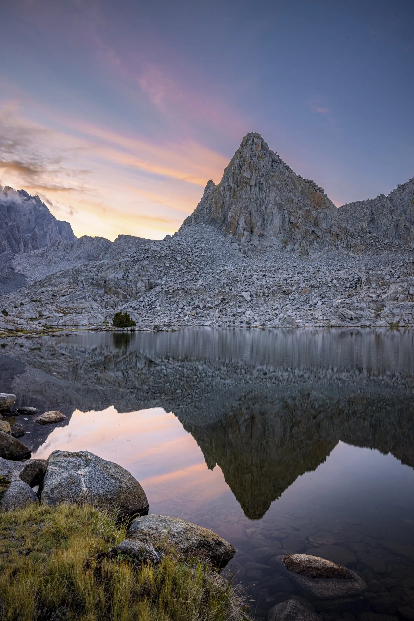 A mountain landscape at sunset with a rugged peak reflected in a calm lake, surrounded by rocky terrain and some grass in the foreground.