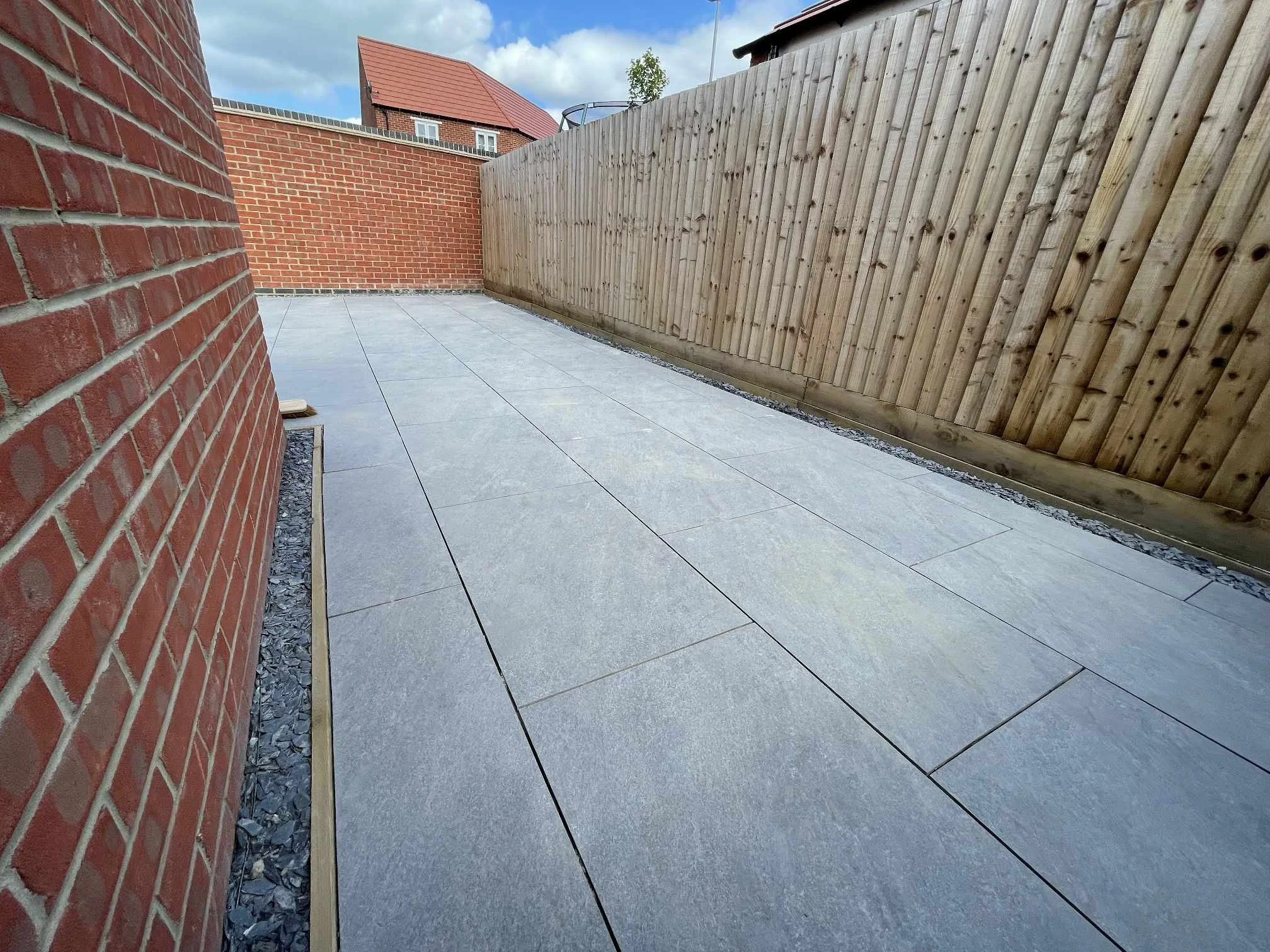 A landscaped outdoor area with large Gray porcelain tiles, bordered by a red brick wall on the left and a wooden fence on the right, under a partly cloudy sky.