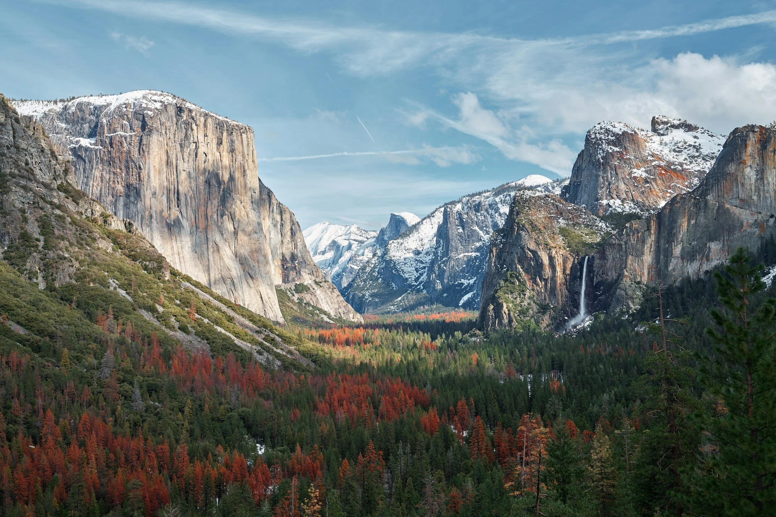 A scenic view of Yosemite National Park with snow-capped mountains, dense pine forests with some trees turning red, and a waterfall on the right side under a partly cloudy sky.