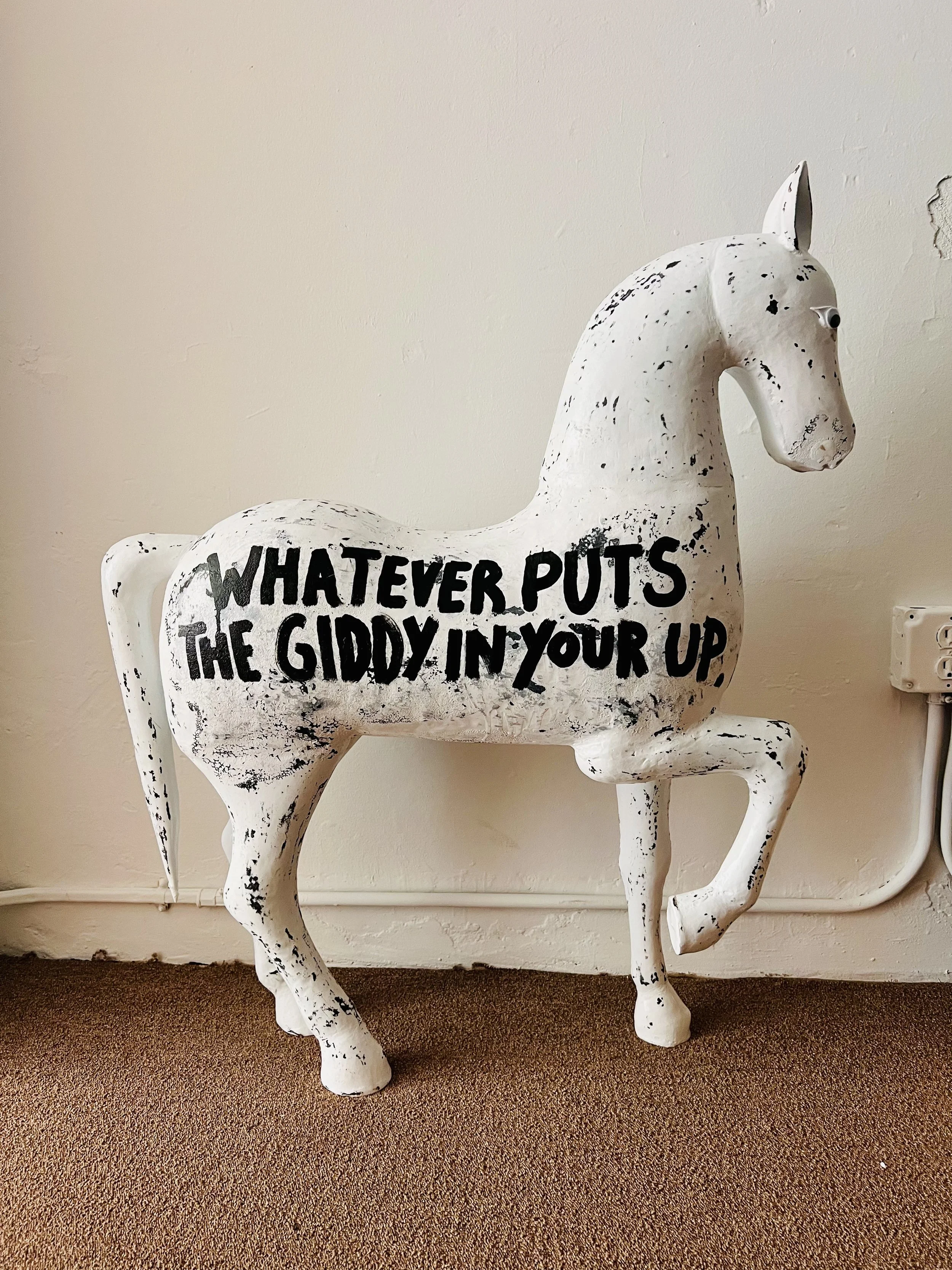 White decorative horse sculpture with black lettering that says 'Whatever puts the giddy in your up' on its side, placed against a beige wall and carpeted floor.