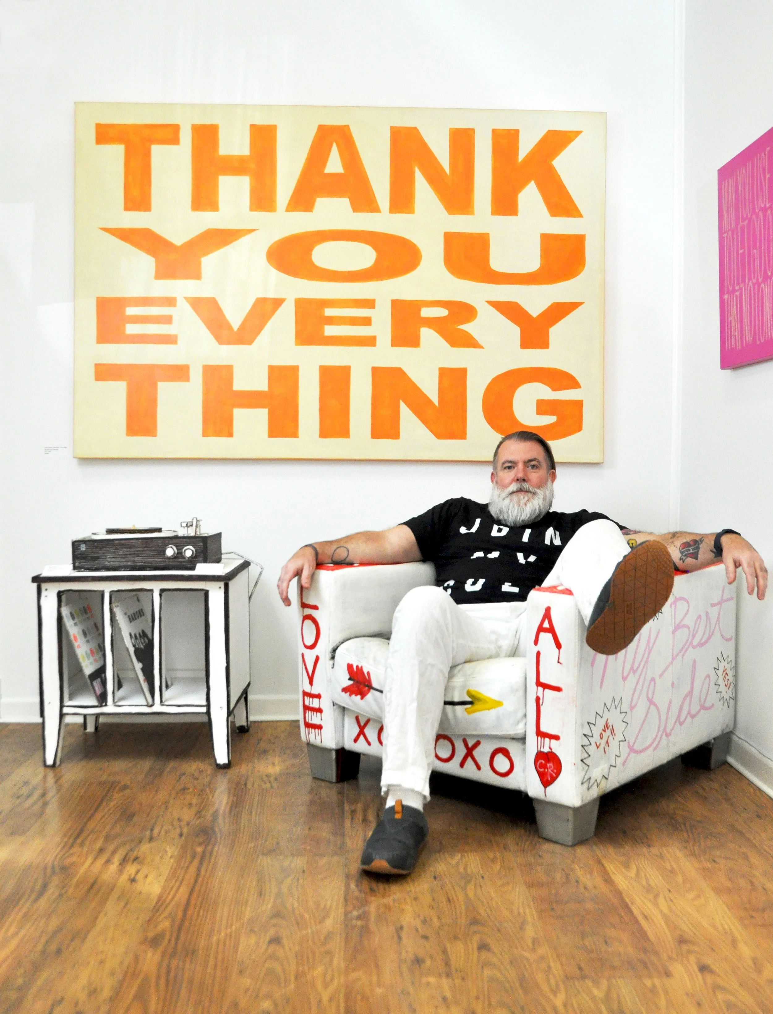 A man with a white beard sitting on a decorated white couch in an art gallery, with a large yellow and orange sign behind him that says 'Thank You Everything'. The couch has words and symbols painted on it, and there is a small black table with a stereo system next to him.