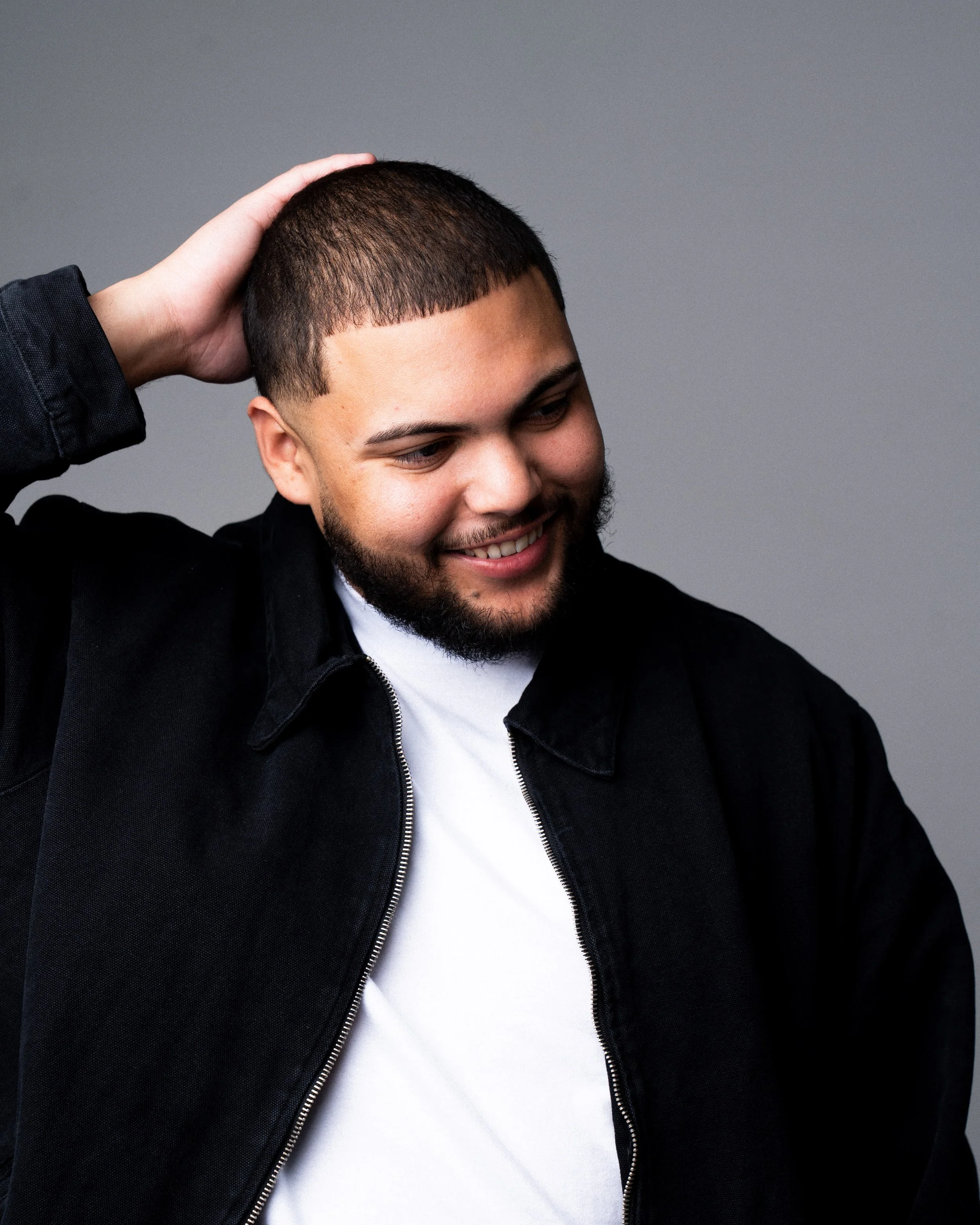 A young man with a beard smiling, wearing a black jacket and white shirt, with one hand on his head against a grey background.