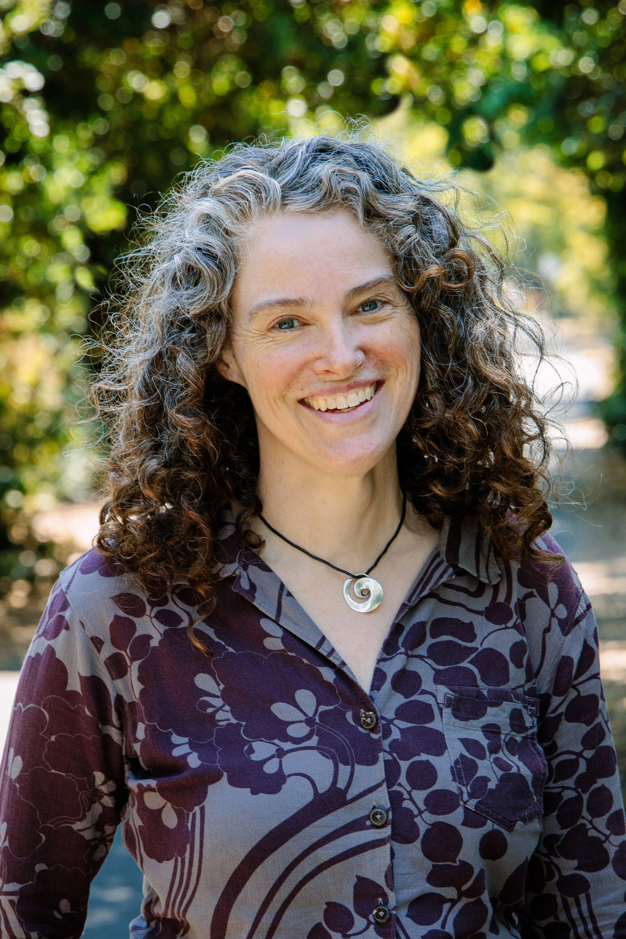 This is a photograph of Julianne Simpson smiling with trees and a path in the background. Julianne has curly hair and is wearing a purple plant patterned shirt and is wearing a round silver pendant.