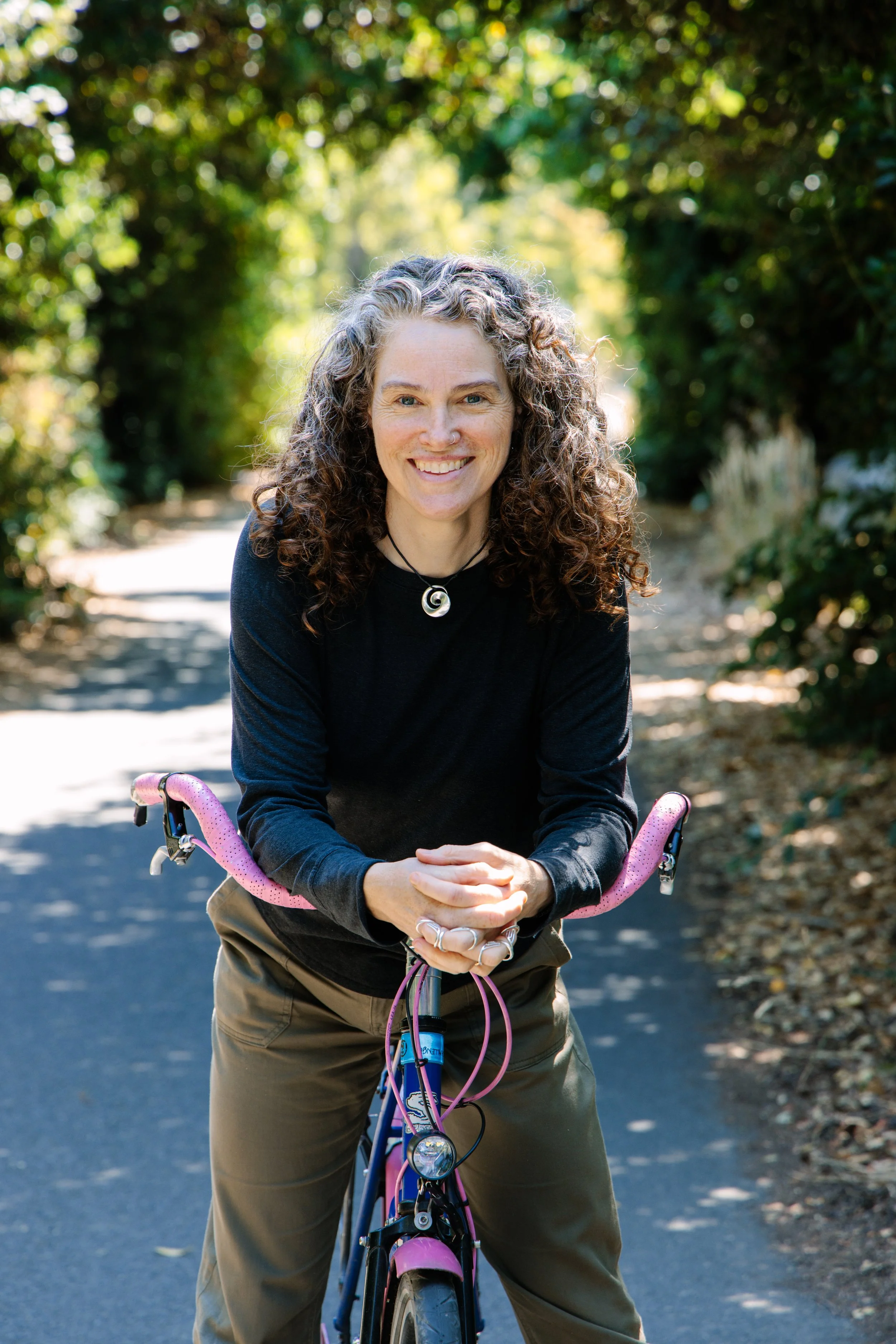 This is a photograph of Julianne Simpson straddling a bicycle, leaning over the handlebars. Julianne has curly brown and gray hair. They are wearing a black shirt and a round silver pendant. They look happy.
