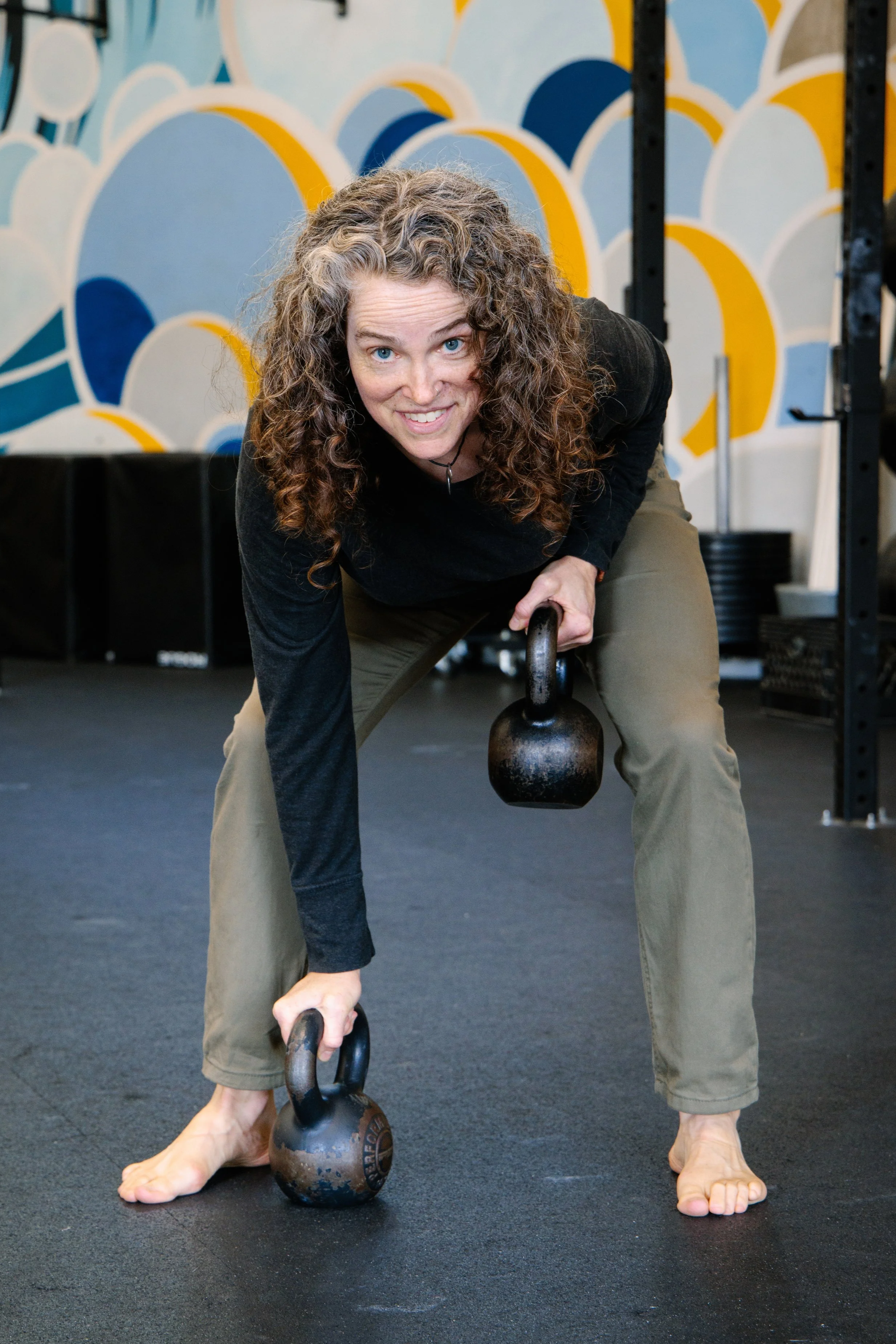 This is a photograph of Julianne Simpson bent forward with a kettlebell in each hand, one on the floor, one at her side. Julianne is smiling.