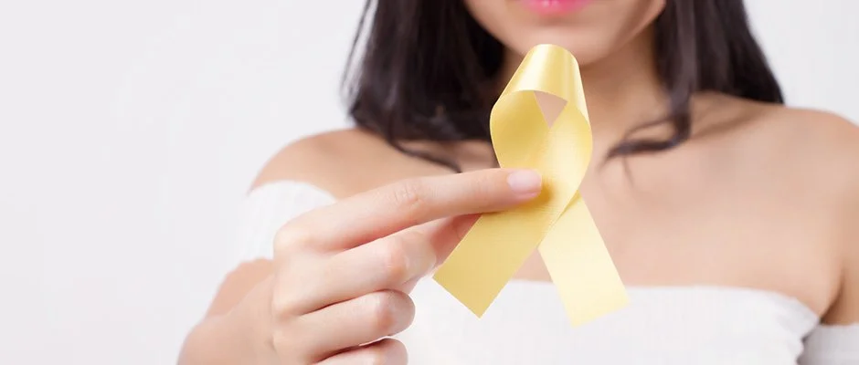 This is a photograph of a femme-appearing person holding a yellow ribbon, the symbol of endometriosis. The photo shows a person’s hand, shoulders, collarbones, chin, and long straight dark hair. They are wearing a white sleeveless top.